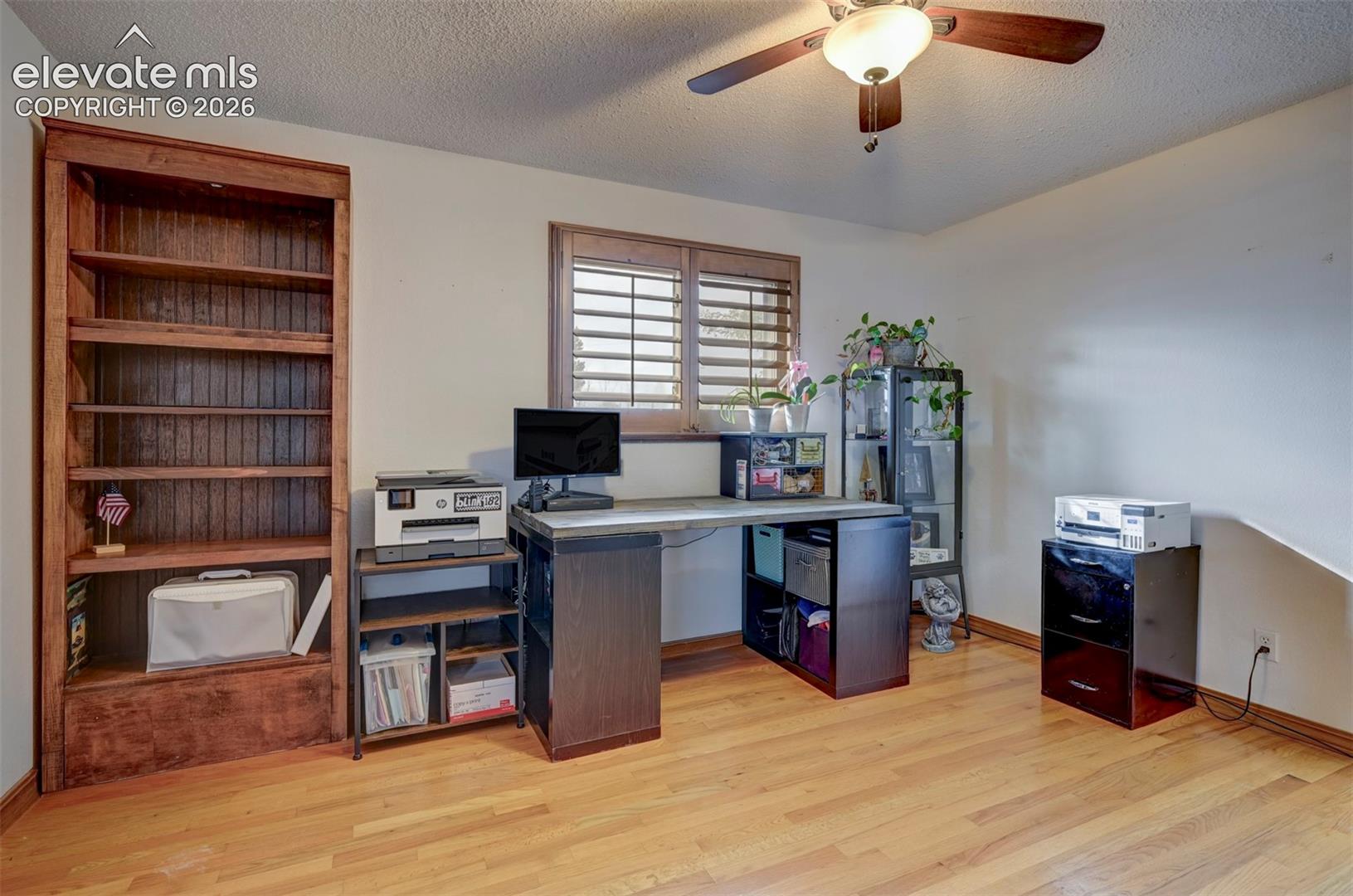 Hardwood flooring, wood-finish built-in shelving, window with plantation shutters, ceiling fan with integrated lighting