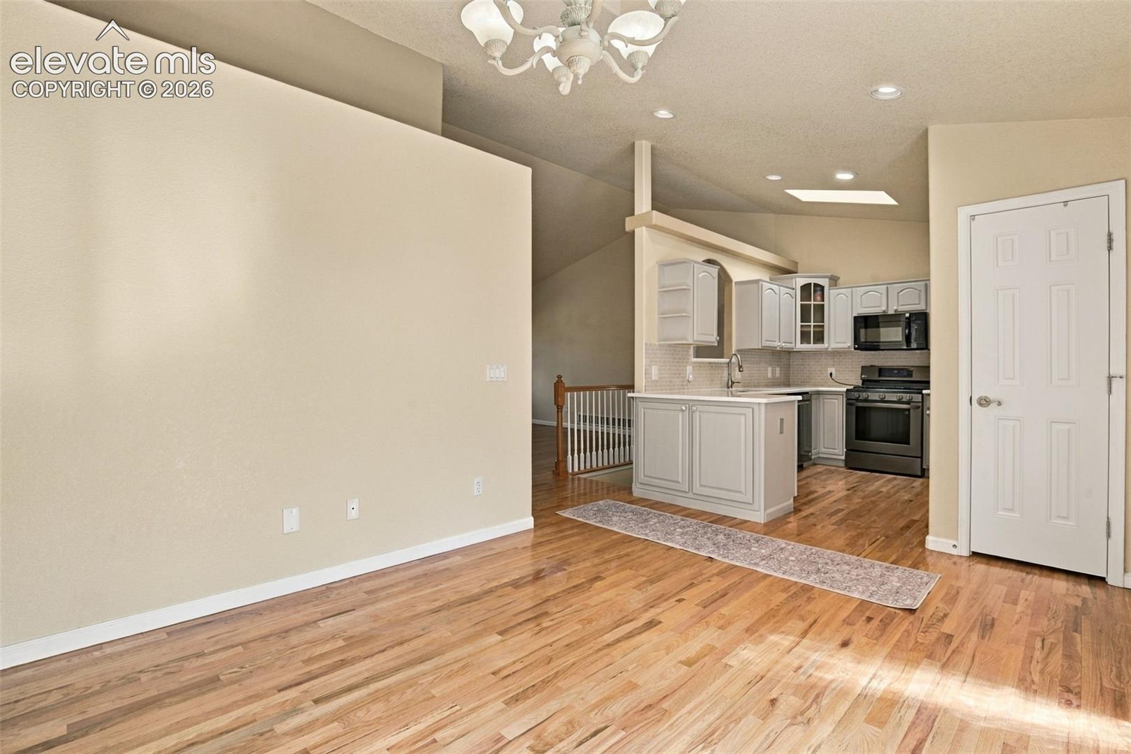 Wood-finish flooring throughout, kitchen featuring light-toned cabinetry, stainless steel appliances, a skylight, and vaulted ceilings with a decorative chandelier