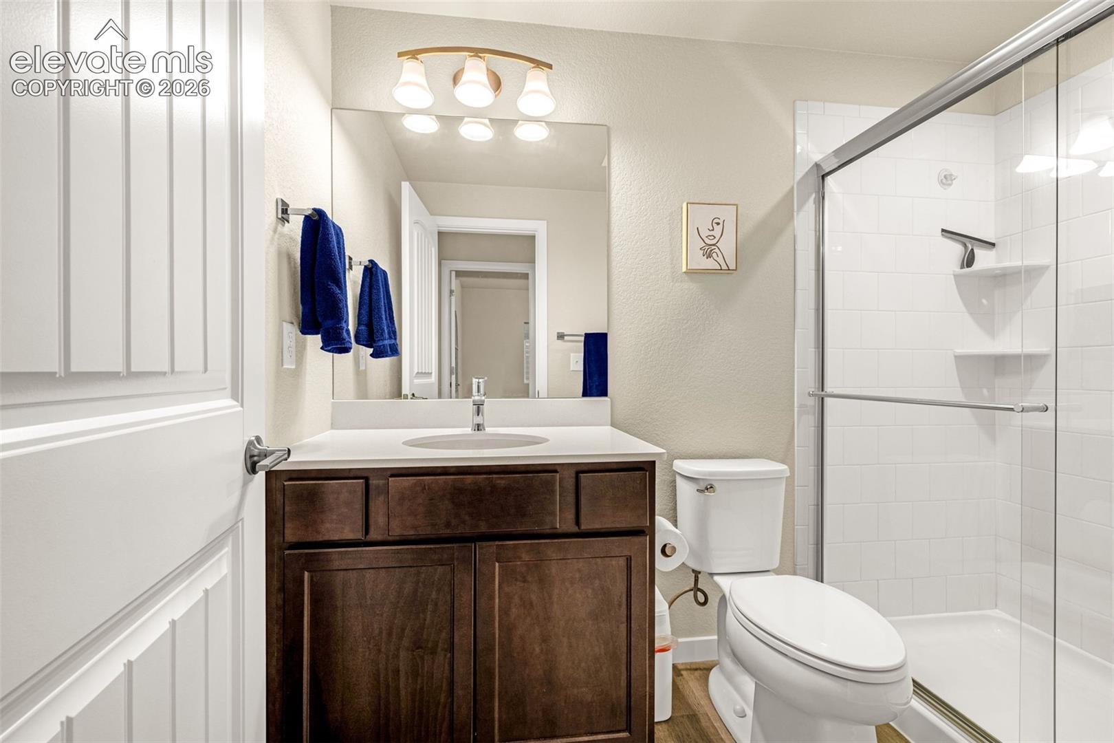 Bathroom featuring a dark wood-finish vanity with a light-toned countertop, integrated basin, frameless mirror, and a glass-enclosed shower with white tiled surround and built-in shelving