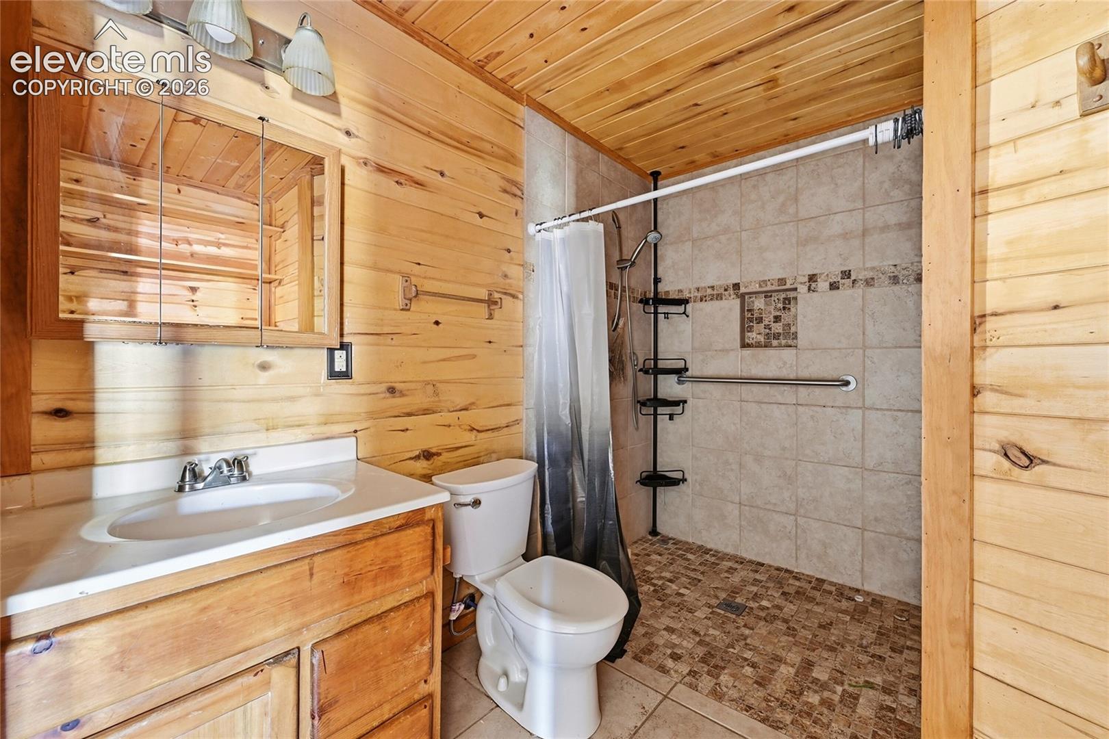 Wood-paneled walls and ceiling, vanity with integrated sink, medicine cabinet with mirrored door, tiled shower enclosure with mosaic accent, and pebble-style shower flooring