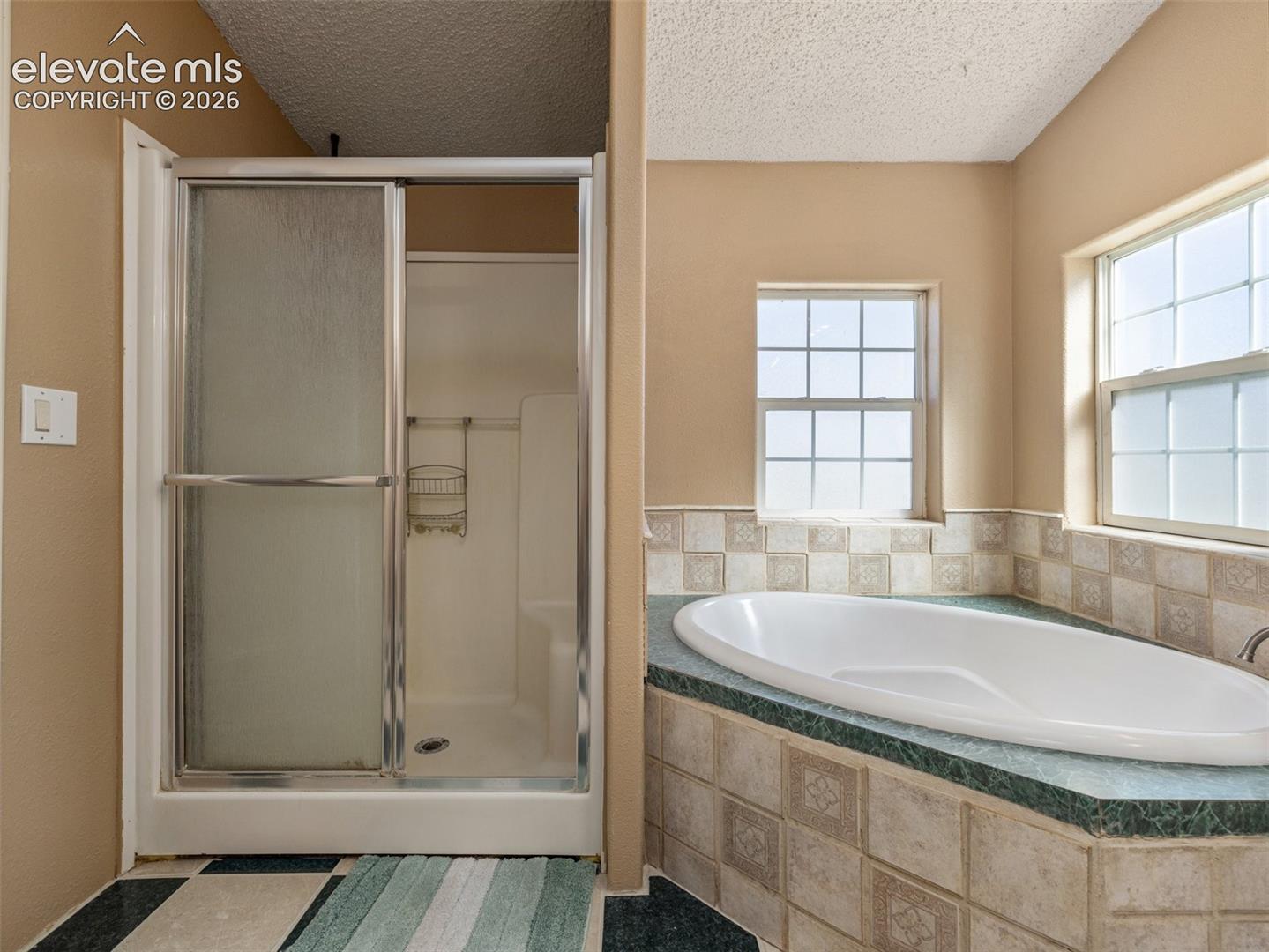 Corner soaking tub featuring a tiled surround and deck, separate shower stall with chrome-framed glass enclosure, and dual windows
