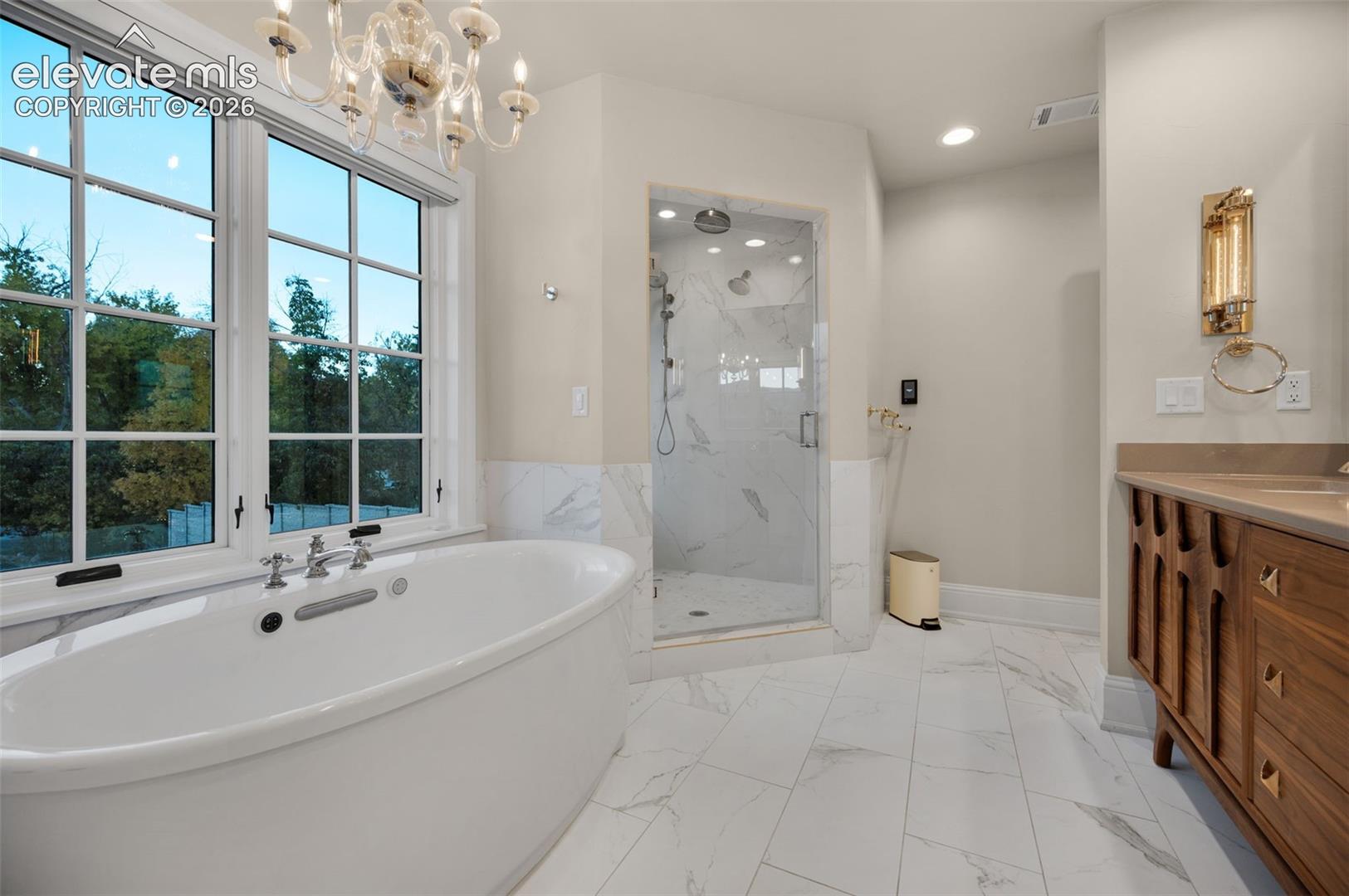 Freestanding soaking tub, walk-in glass-enclosed shower, stone-finish tile flooring, wooden cabinetry, and crystal-style chandelier lighting