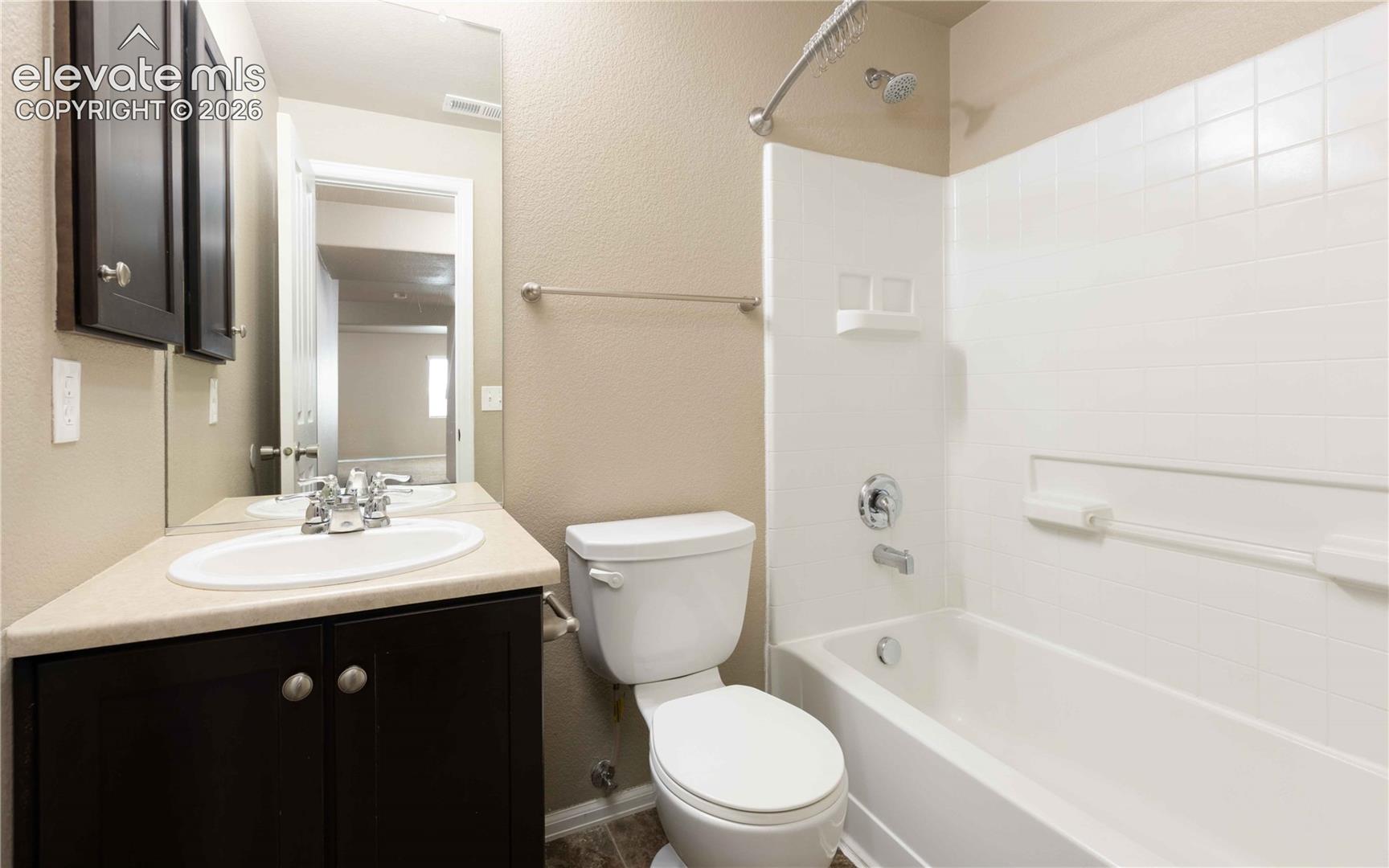 Dark-finish cabinetry with a neutral countertop, undermount sink, wall-spanning mirror, and a tub-shower combination with white tile surround