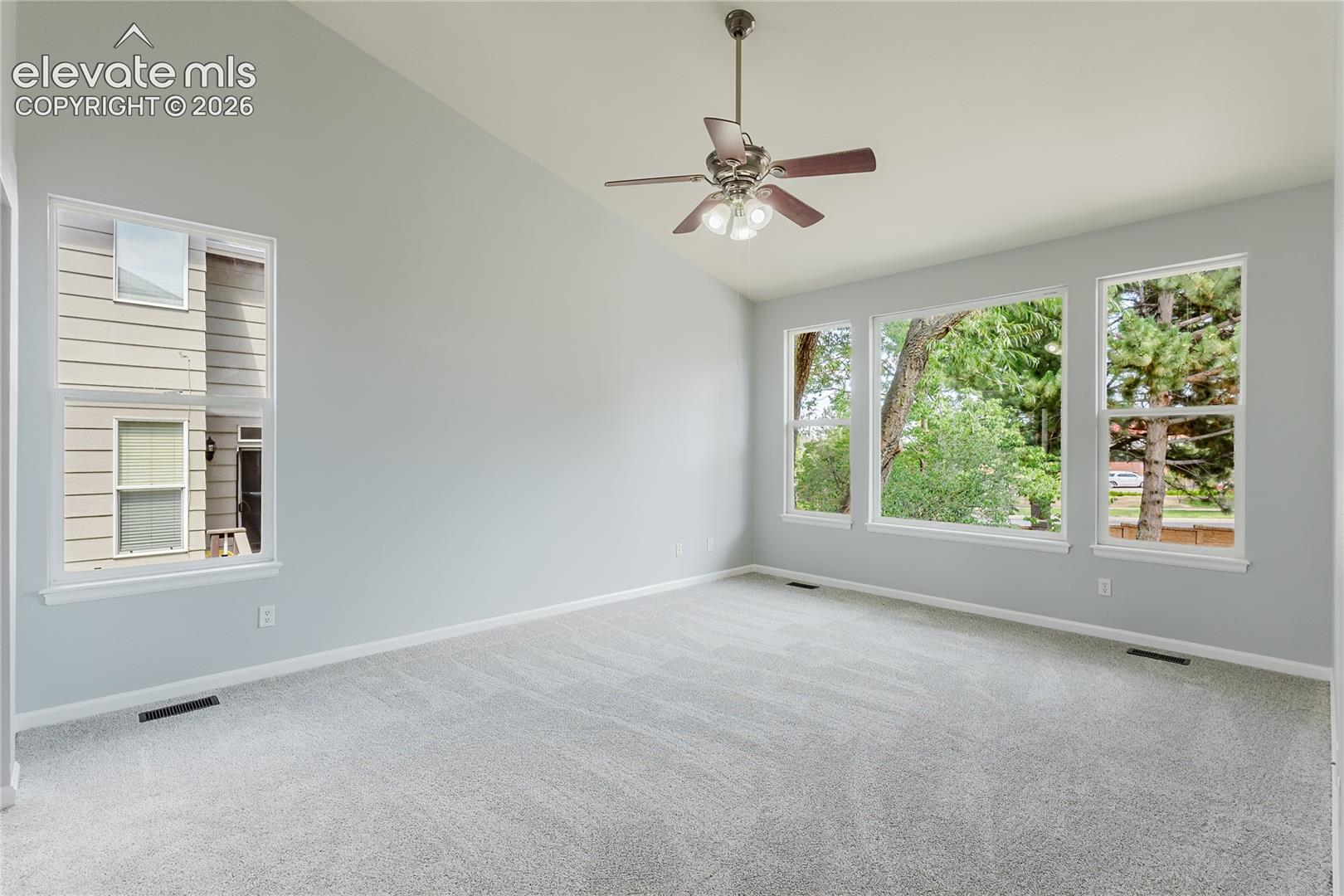 Vaulted ceiling with a ceiling fan light fixture, light gray carpet, multiple windows with white trim, and light gray walls