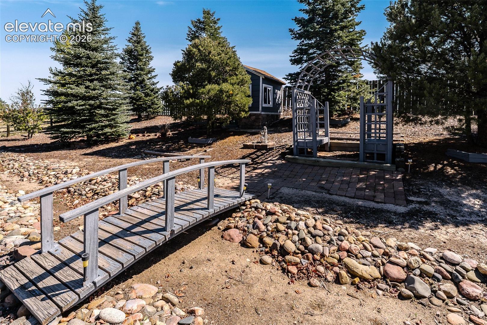 Backyard bridge, arched trellis, and garden shed behind the trees.