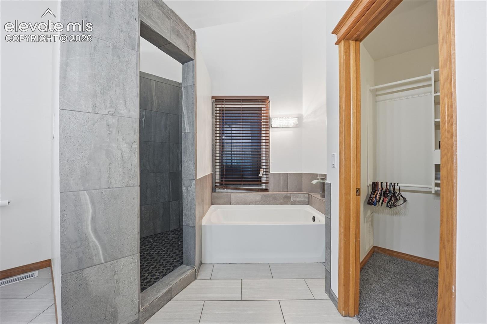 Tiled shower enclosure, soaking tub, window with blinds, and built-in closet organization