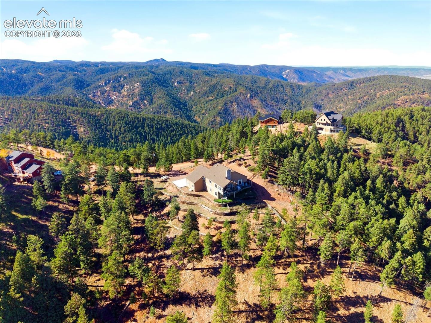 Elevated mountain landscape featuring a sprawling residence with a multi-level stone retaining wall, paved driveway, and expansive forest surroundings