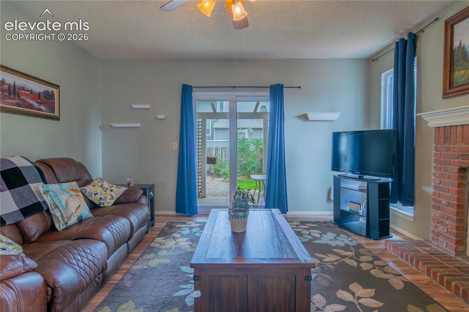Living area with wood-finish flooring, a brick fireplace, and a ceiling fan with integrated lighting