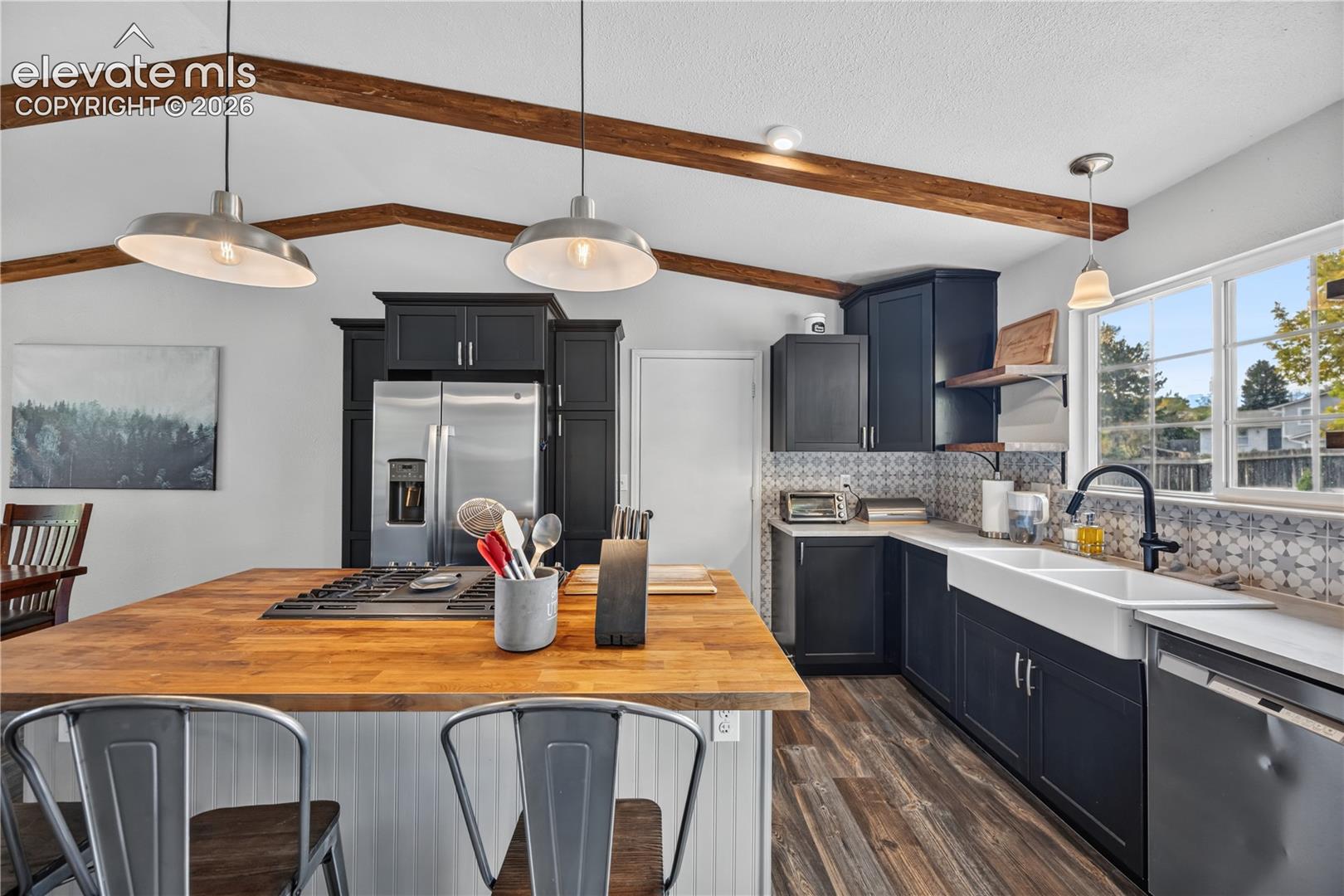 Kitchen featuring exposed vaulted ceiling beams, dark cabinetry, butcher block island countertop, apron-front farmhouse sink, and patterned backsplash
