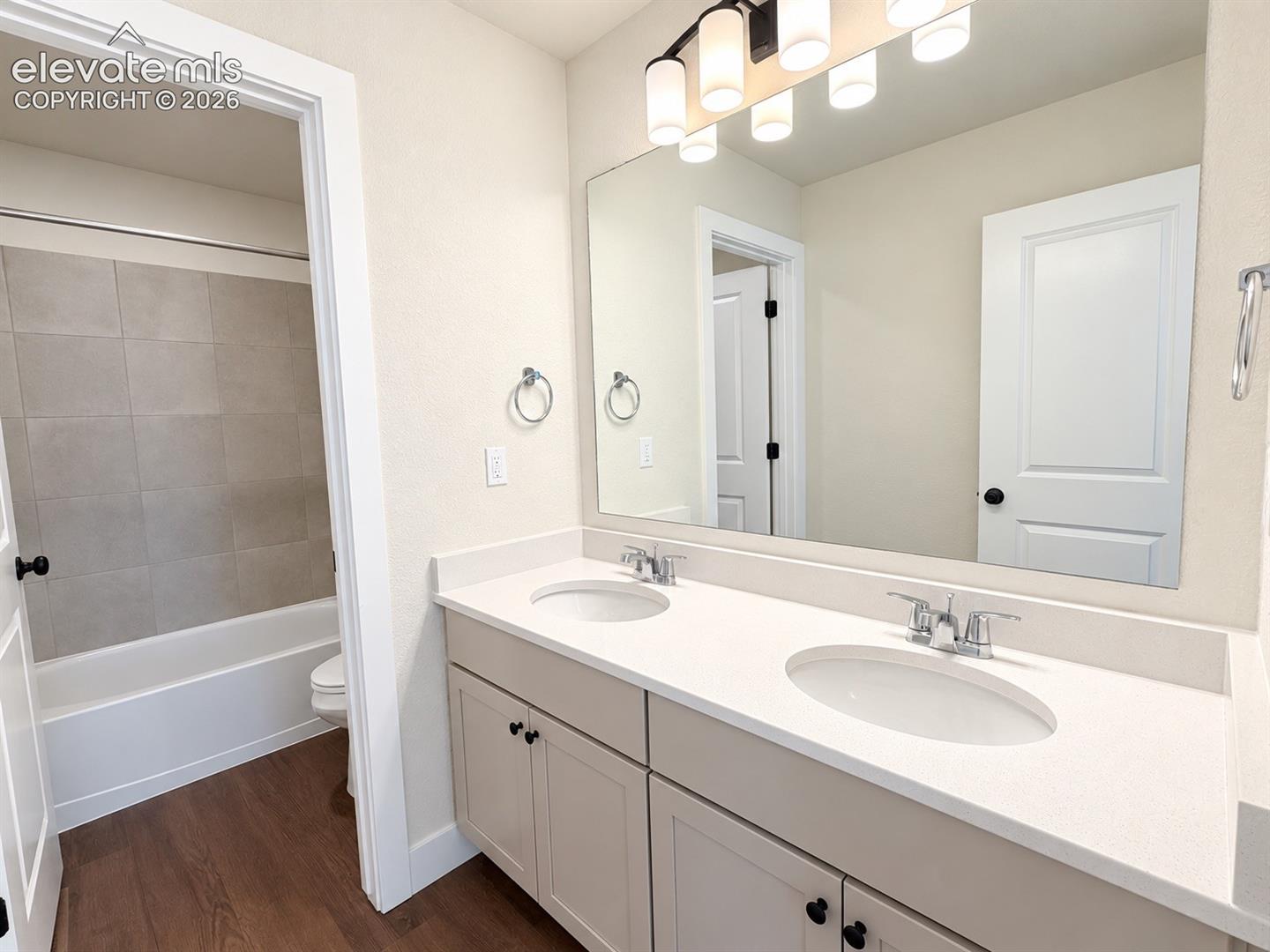 Dual vanity with white countertops and undermount sinks, wood-finish flooring, white cabinetry with black hardware, and a tub with tiled surround