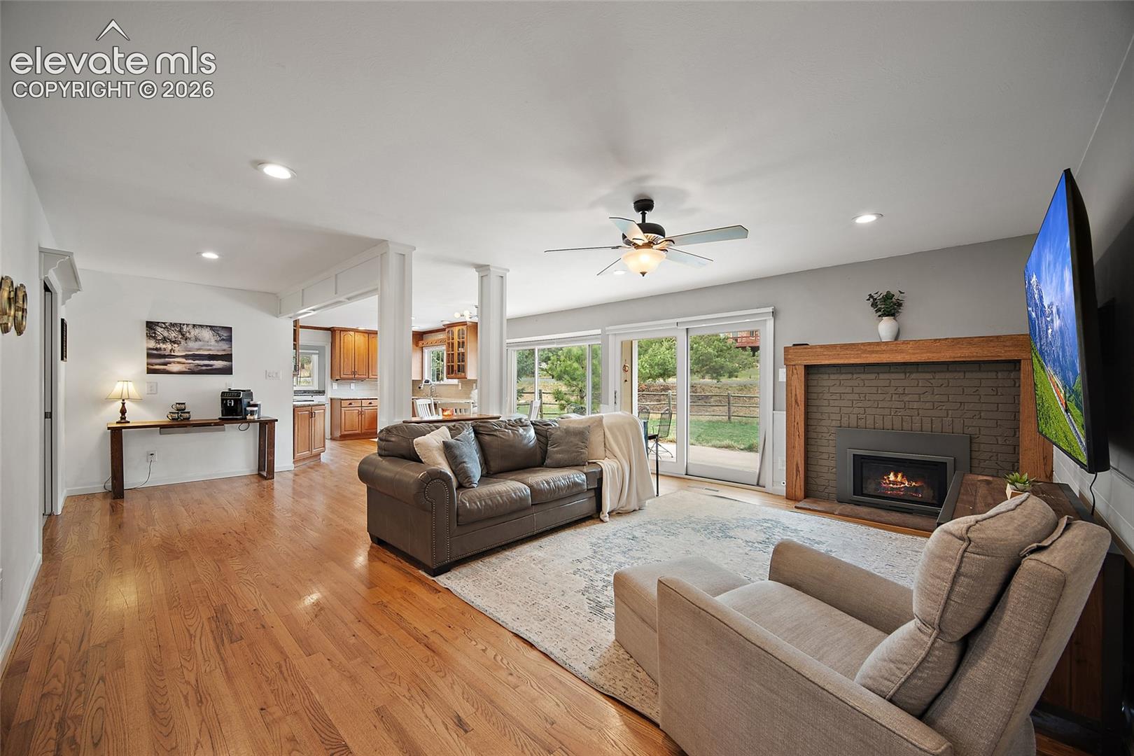 Open-concept interior featuring wood-finish flooring, a painted brick fireplace with a natural wood mantel, recessed lighting, and sliding glass doors