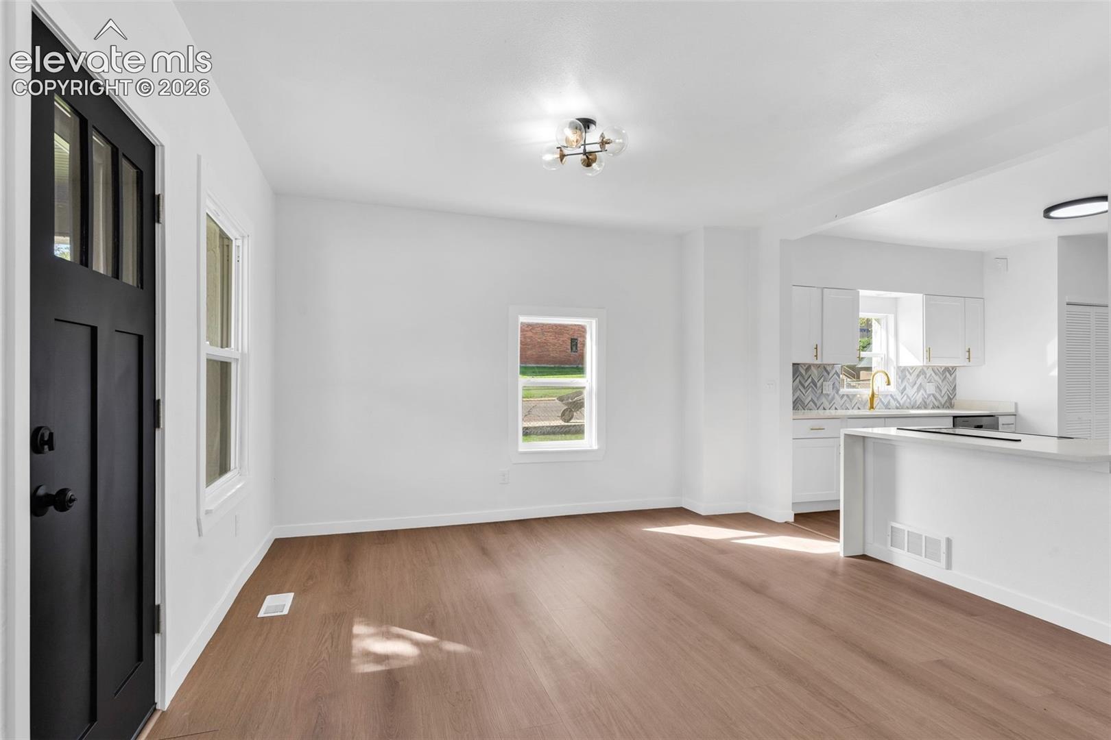 Open-concept interior featuring wood-finish flooring, a matte black multi-lite entry door, a multi-arm flush mount light fixture, and white shaker-style cabinetry with a herringbone pattern backsplash
