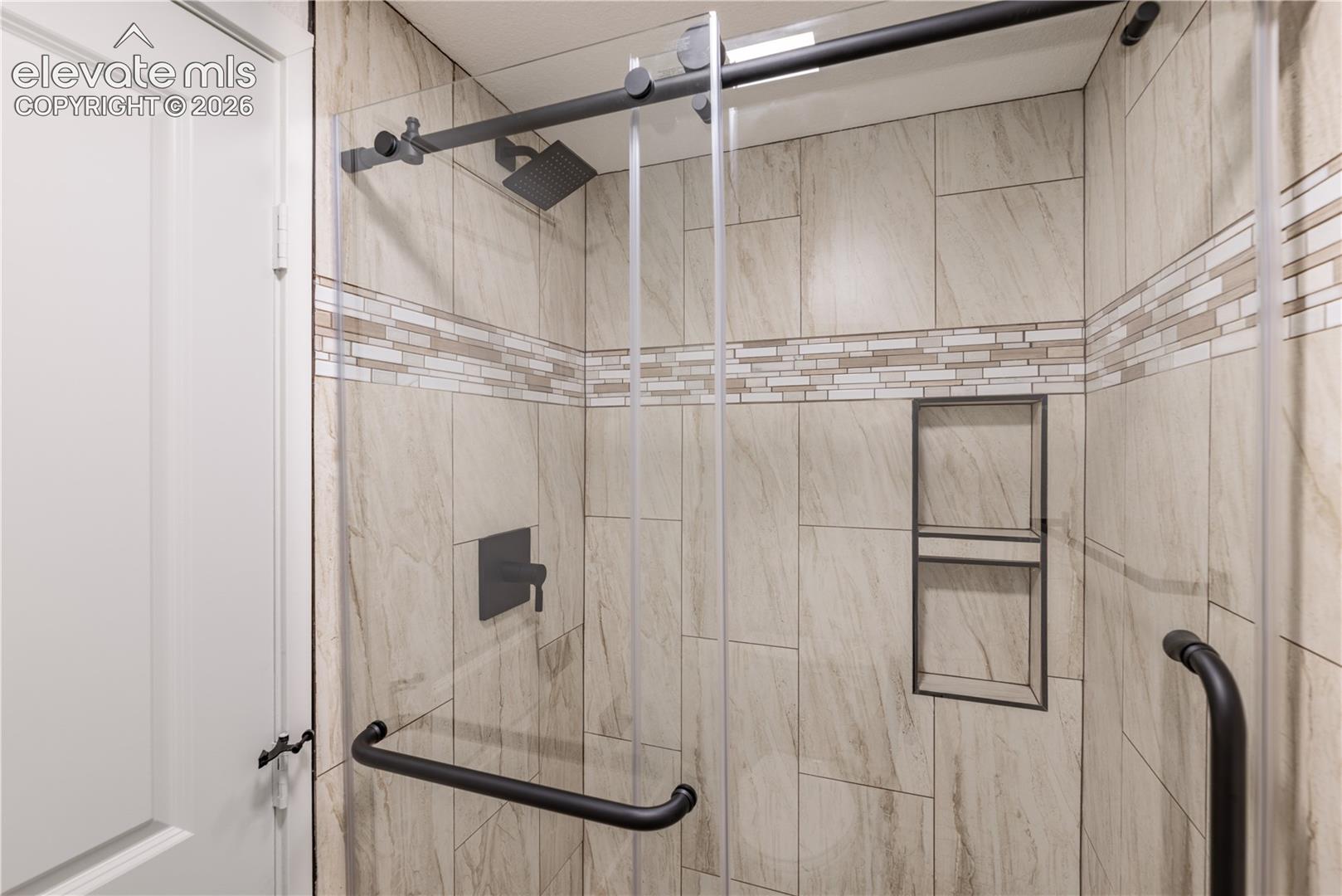 Walk-in shower featuring large-format stone-finish wall tile, decorative horizontal mosaic tile inlay, built-in recessed wall shelving, matte black fixtures, and a frameless glass sliding door