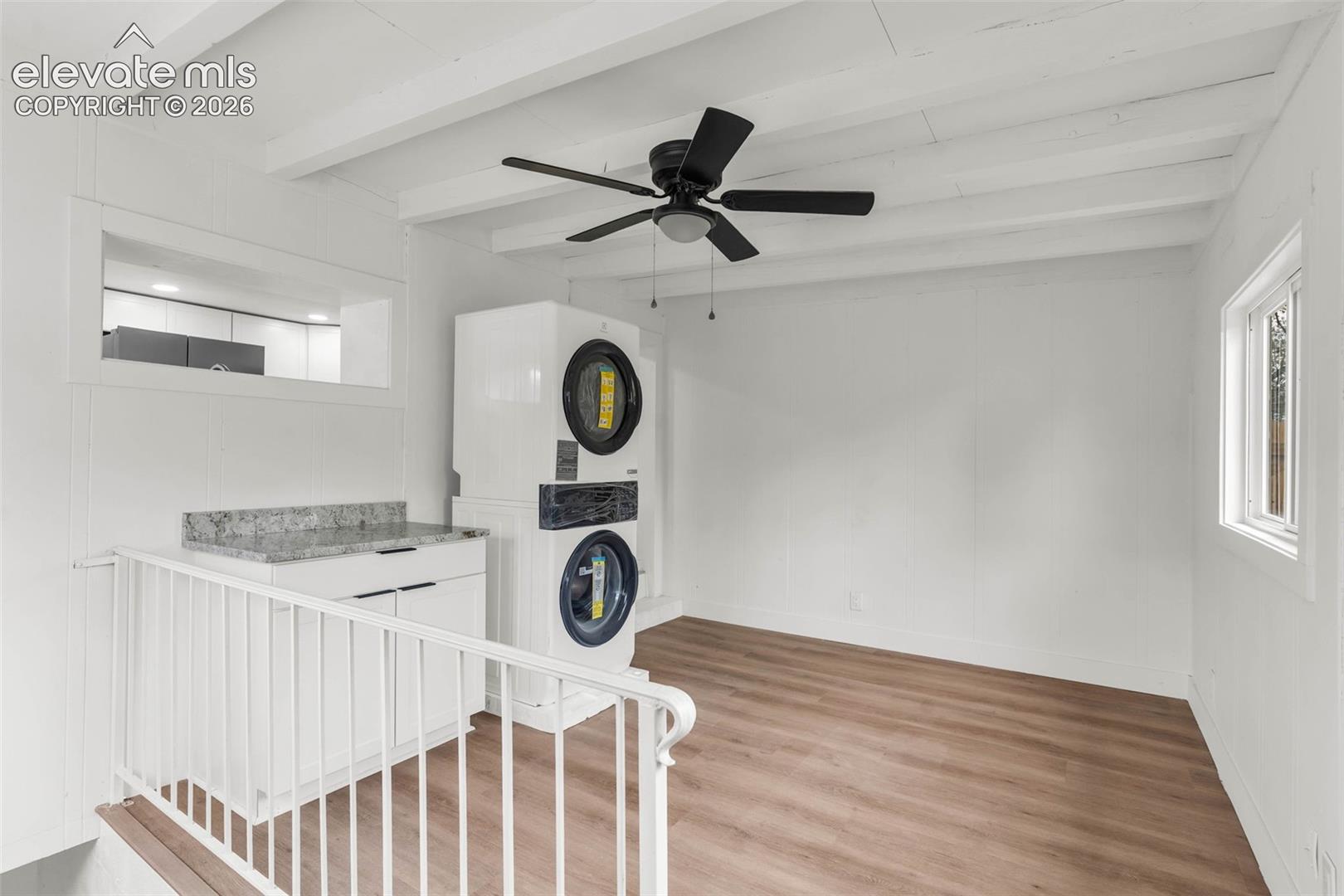 Wood-finish flooring, white exposed ceiling beams, stacked laundry unit, white cabinetry with stone-finish countertop, and black ceiling-mounted fan