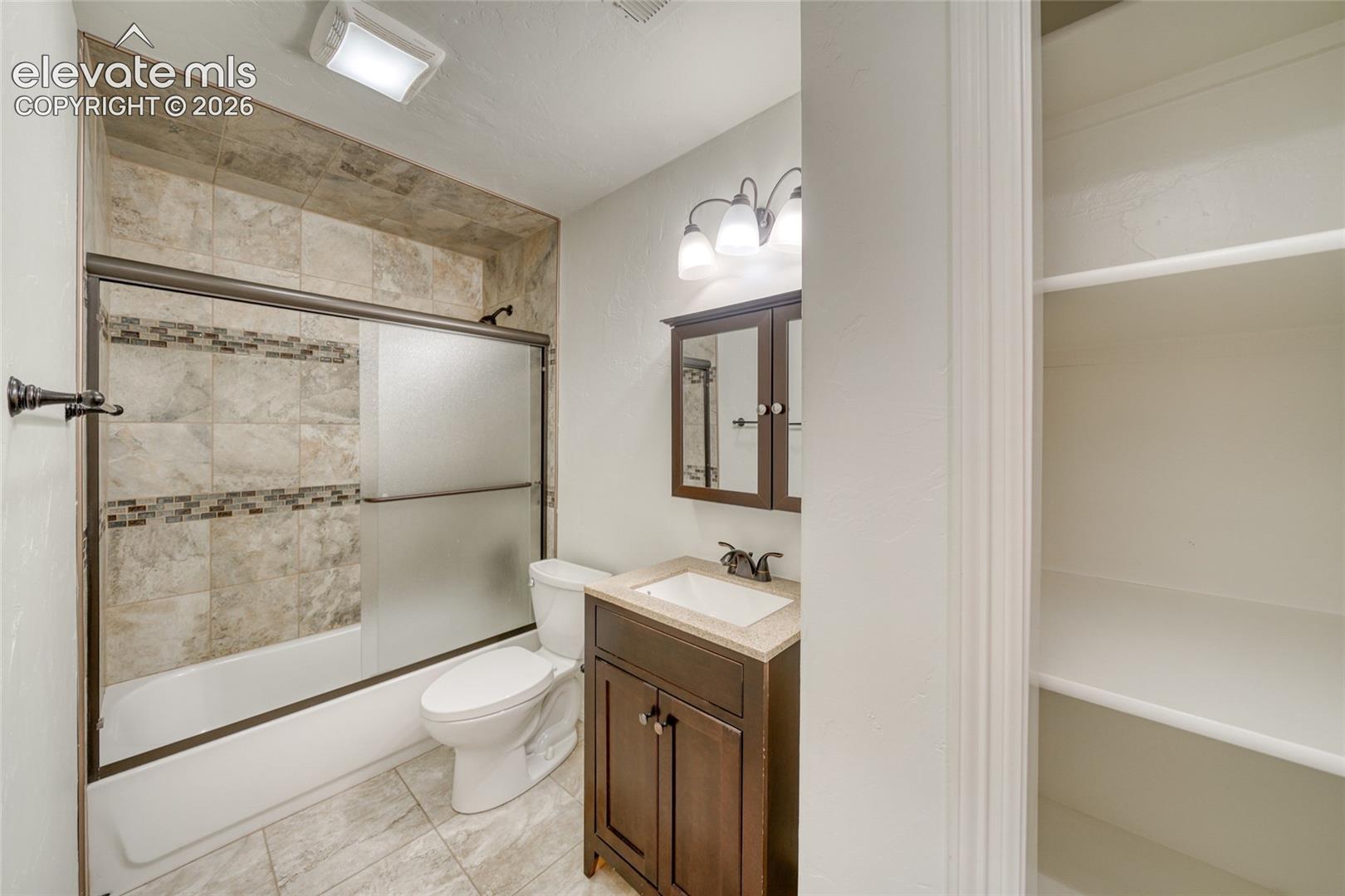 Bathroom featuring a tiled shower surround with decorative accent, a bathtub with sliding frosted glass doors, a single vanity with storage, and a built-in shelving unit