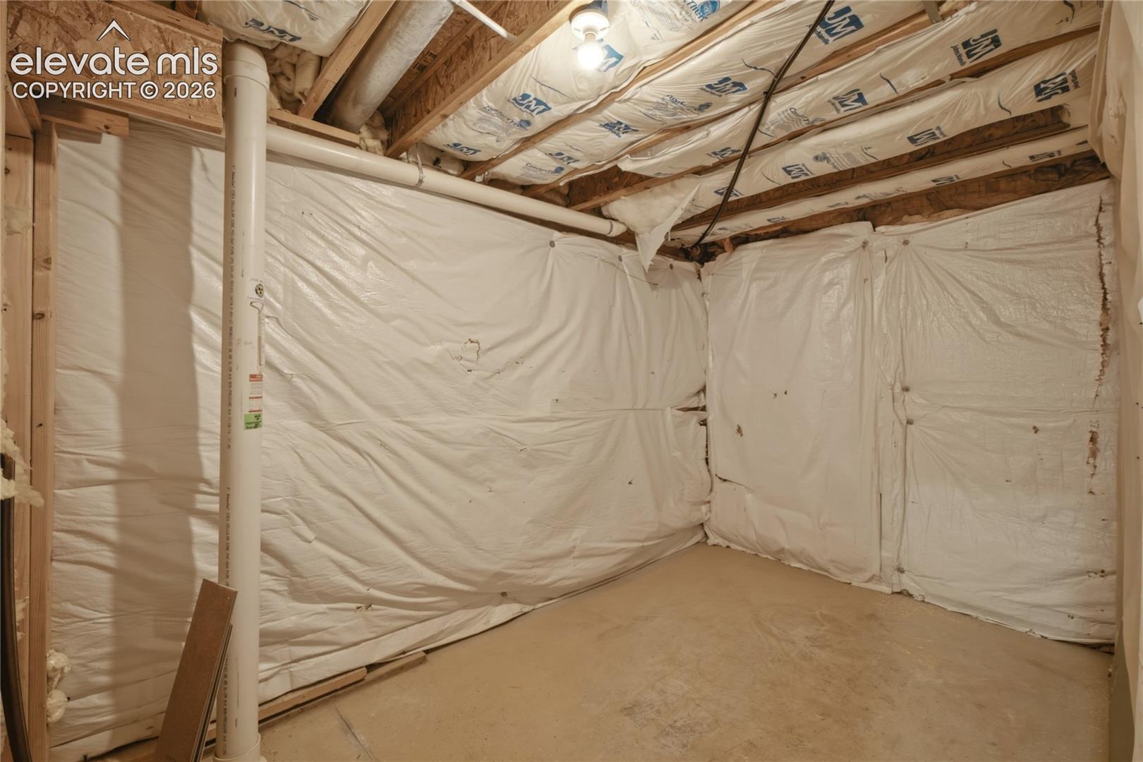 Unfinished basement space featuring exposed wood joists and subflooring, white plastic vapor barrier on walls, and a concrete floor