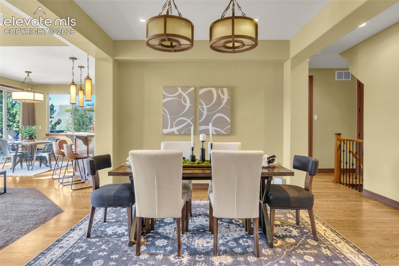 Dining area featuring dual drum-style pendant lights, wood-finish flooring, and a decorative staircase with a wood handrail and iron balusters