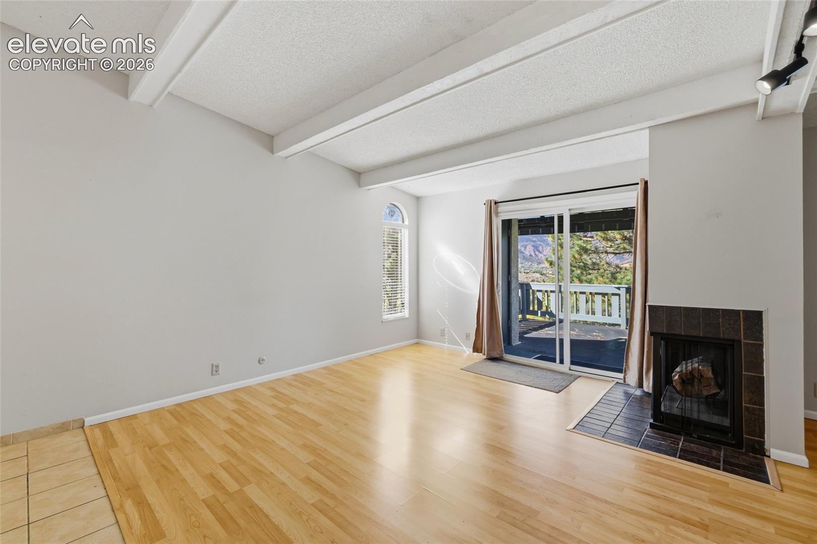 Living area featuring vaulted ceilings with exposed beams, wood-finish flooring, a tile-surround fireplace, and a sliding glass door leading to a balcony