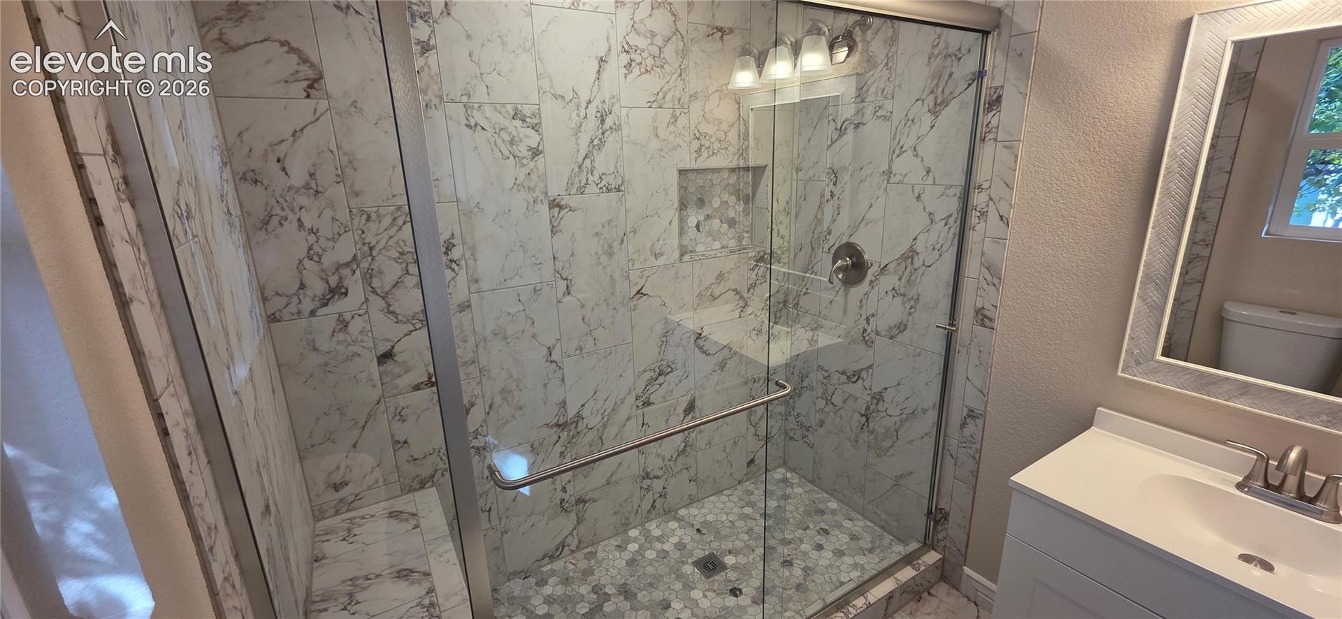 Glass-enclosed shower with marble-style tile surround, mosaic tile shower pan, built-in shower niche, and brushed nickel fixtures