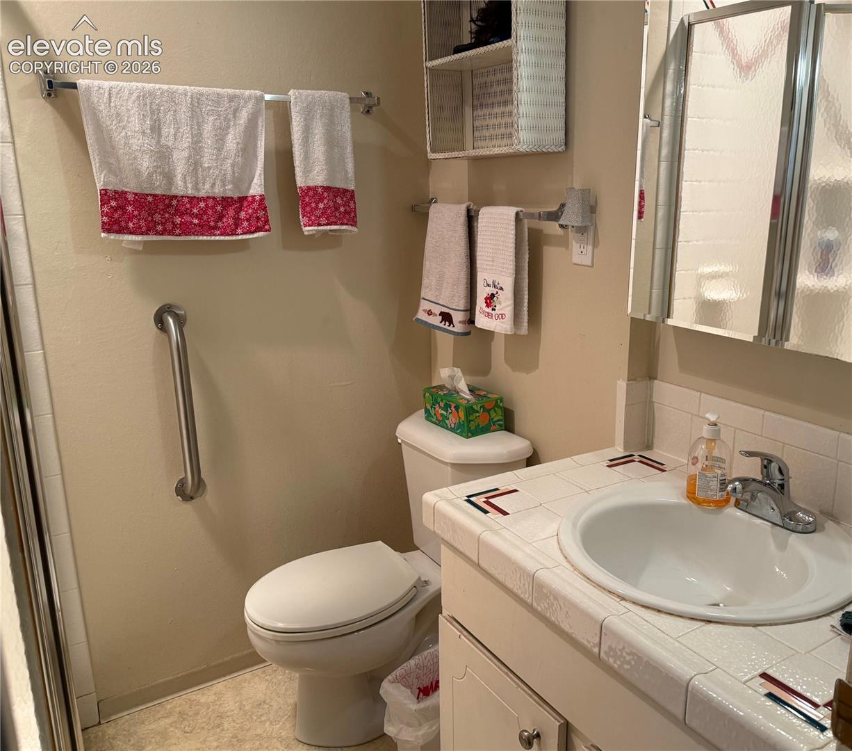 Bathroom featuring a tiled vanity countertop with an integrated basin, a wall-mounted grab bar, a mirrored medicine cabinet, and integrated shelving