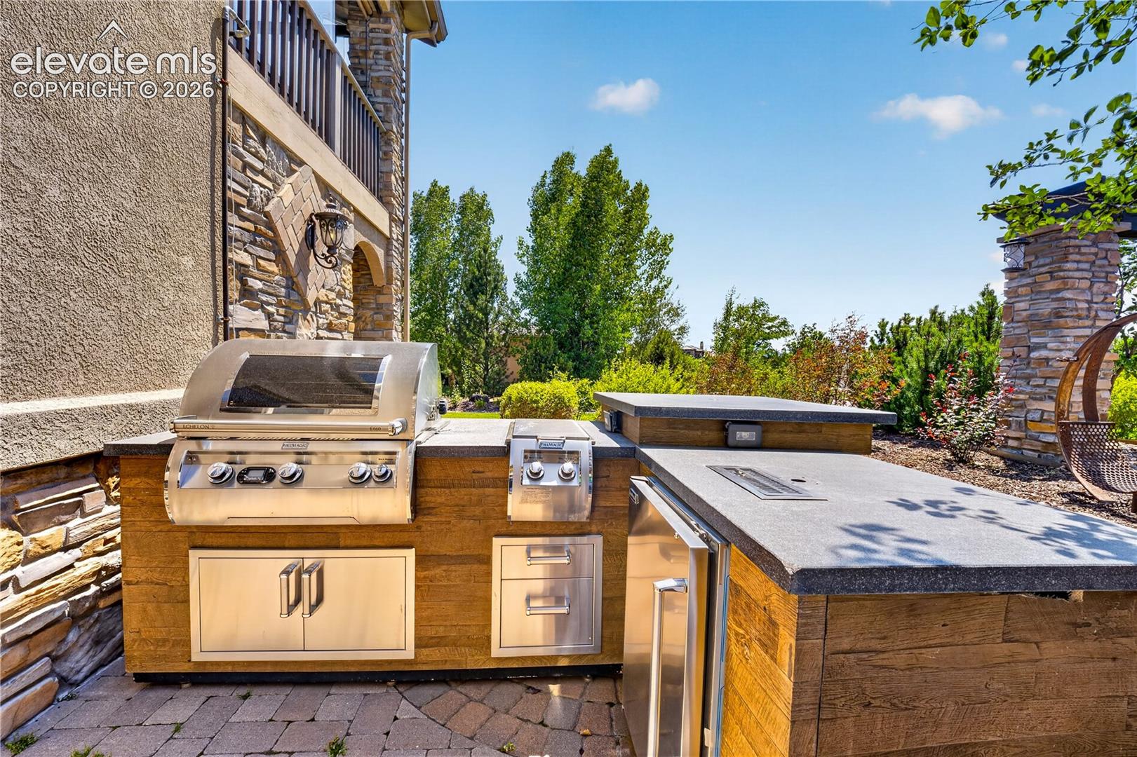 Outdoor kitchen featuring a stainless steel grill and burner, custom wood-paneled base cabinetry, stone countertops, and an integrated refrigerator, situated on a stone paver patio