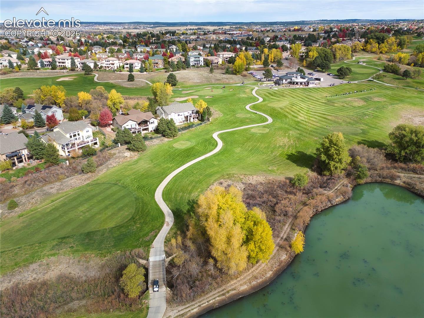 Aerial perspective of a sprawling golf course featuring expansive manicured greens, a paved cart path, a concrete bridge, and a prominent water feature