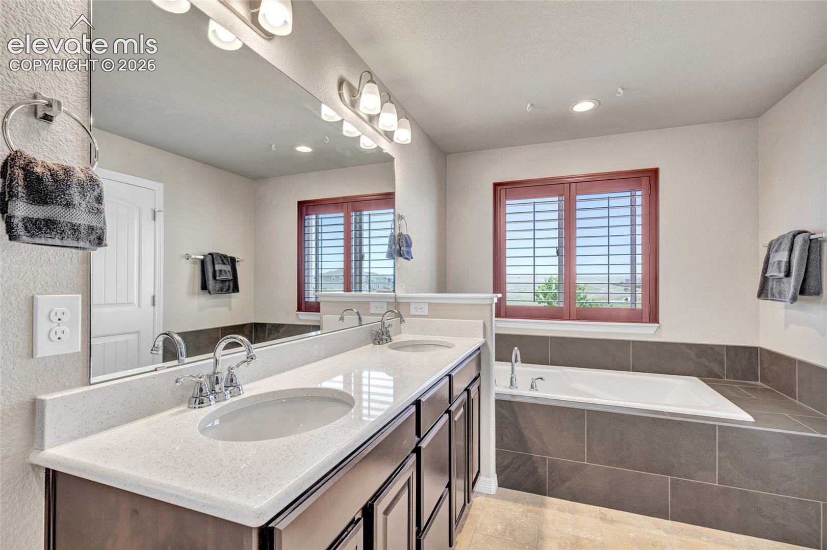 Spacious bathroom featuring a dual vanity with light-toned countertops, chrome fixtures, a large soaking tub with dark tile surround, and plantation shutters