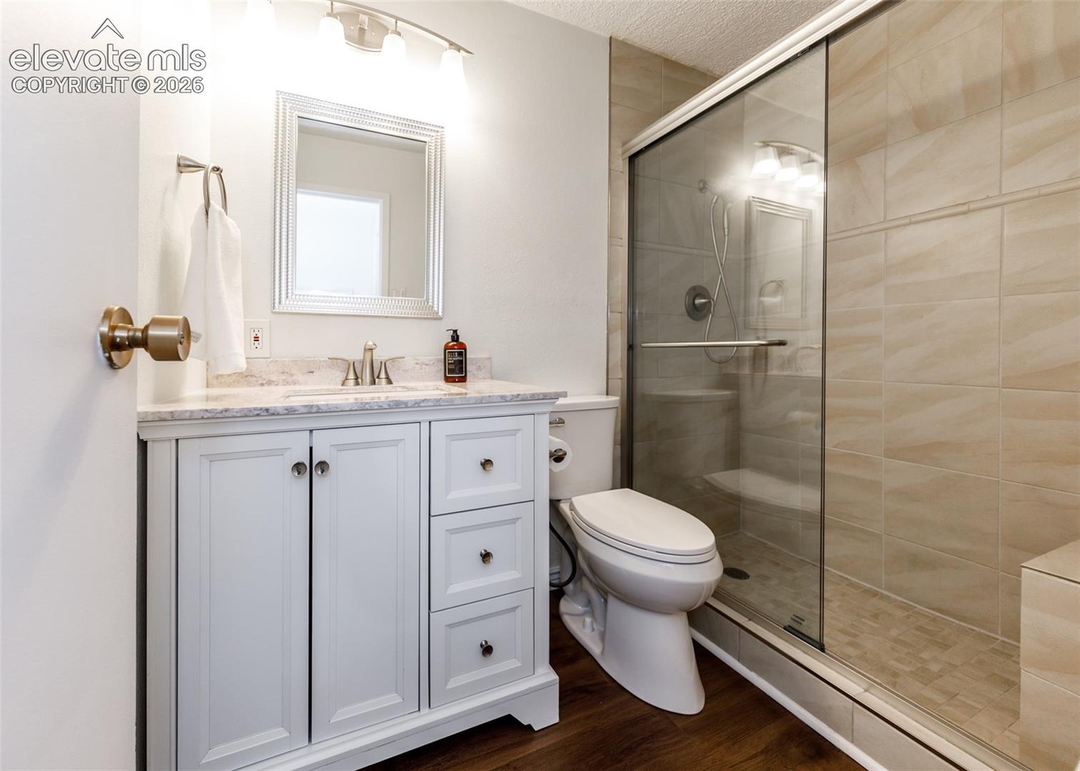 White vanity with stone-composite countertop, integrated sink, shower stall with sliding glass door, tiled shower surround, and wood-finish flooring