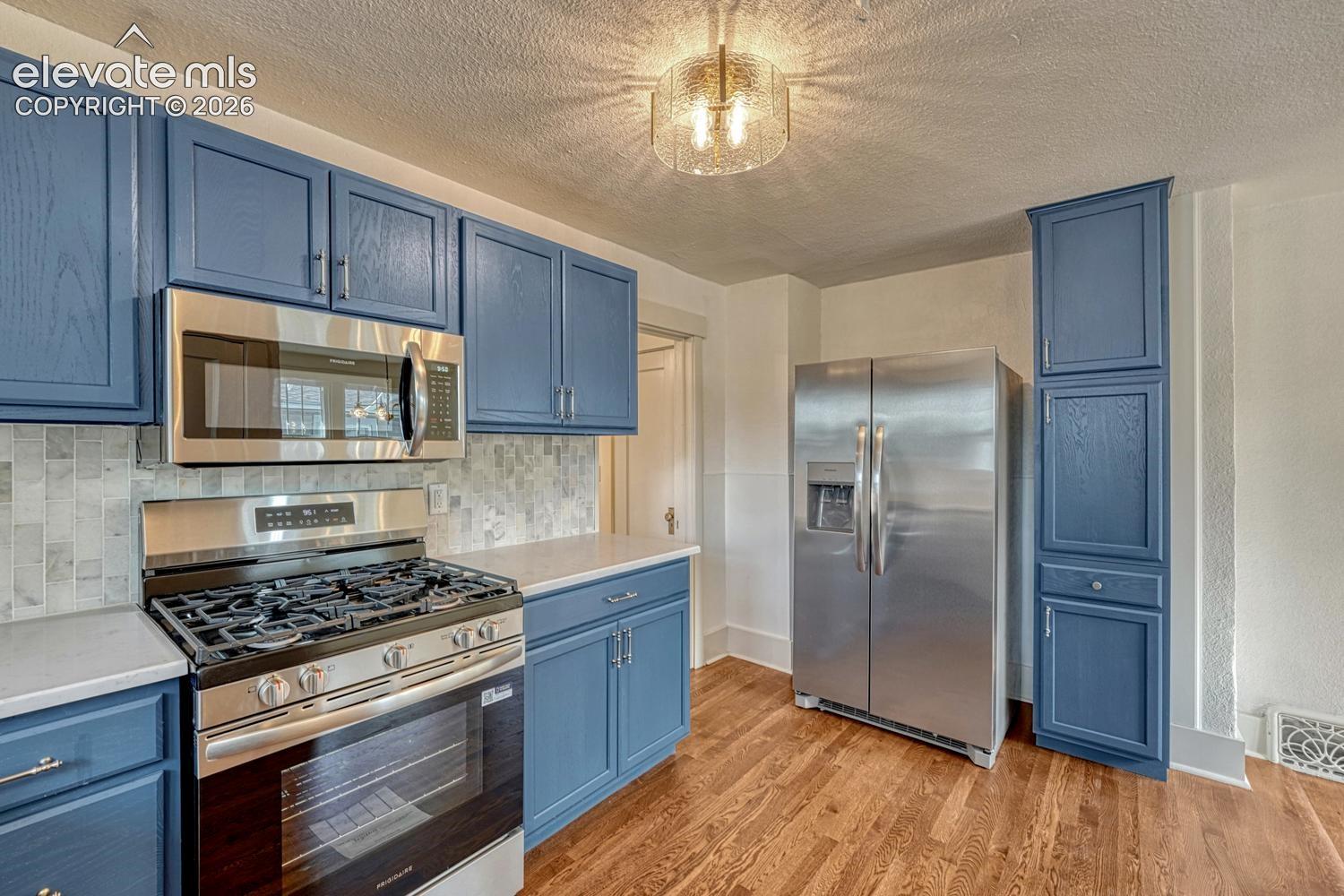 Blue cabinetry with stainless steel appliances, mosaic tile backsplash, light countertops, and wood-finish flooring