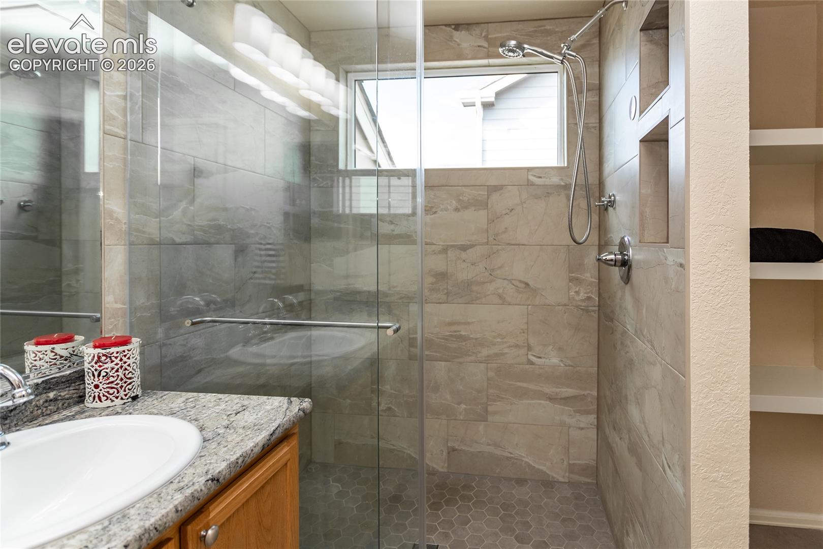 Walk-in shower featuring large-format wall tile, a frameless glass enclosure, a polished chrome rainfall showerhead, integrated tiled shelving, and a natural stone vanity countertop