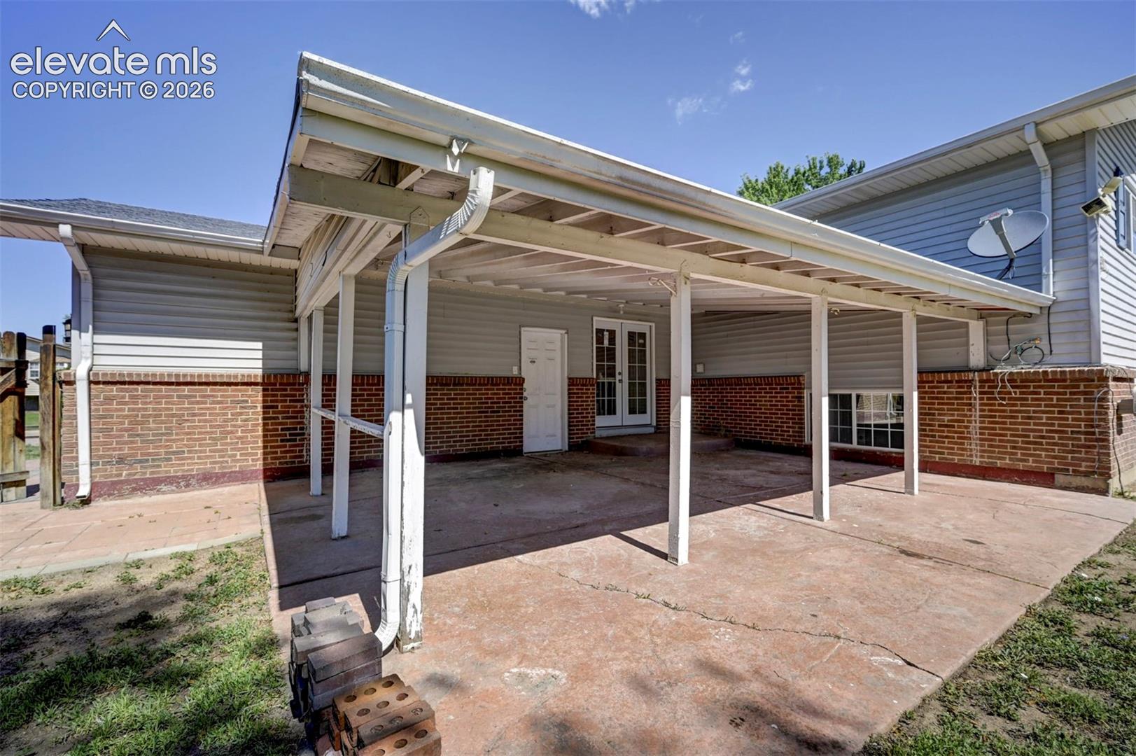 Covered concrete patio featuring white support posts, horizontal siding, brick masonry, and double-door entry