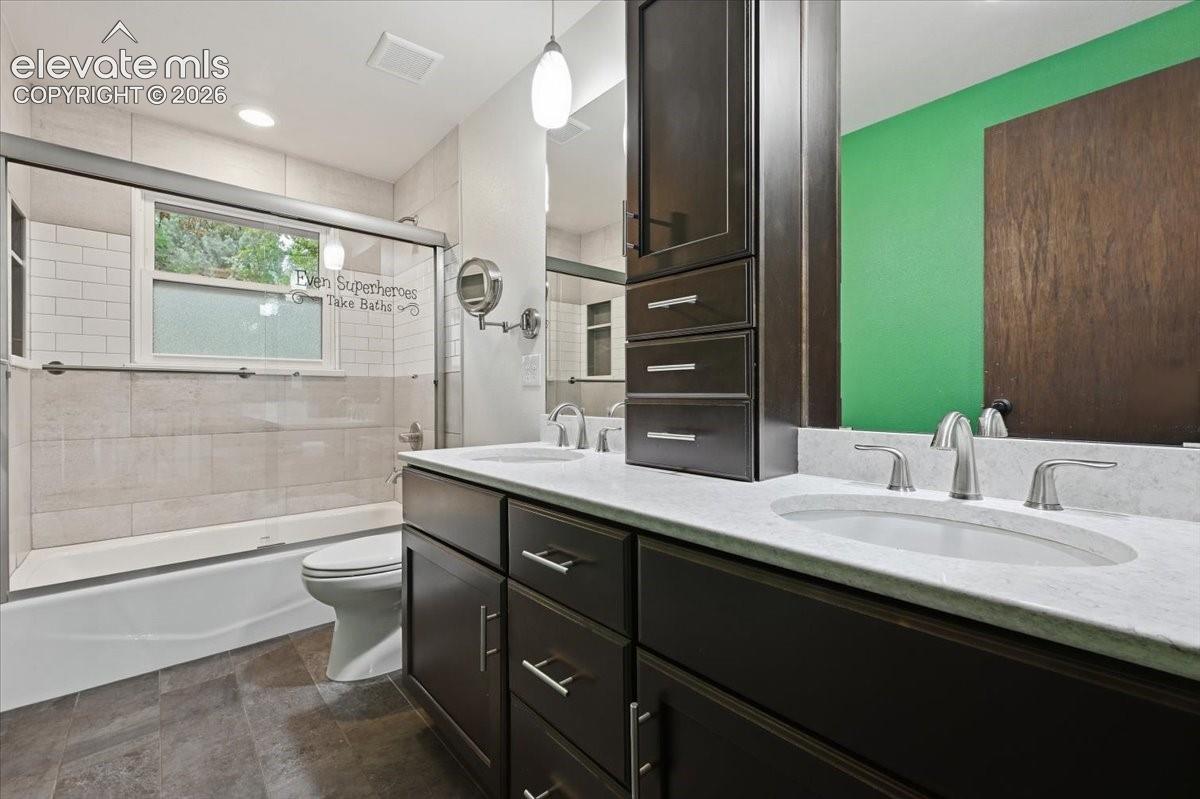 Dual vanity with under-mount sinks and brushed nickel faucets, dark wood-finish cabinetry, built-in storage tower, and a combination tub/shower with glass enclosure. Cabinetry has added electrical for charging inside cabinets