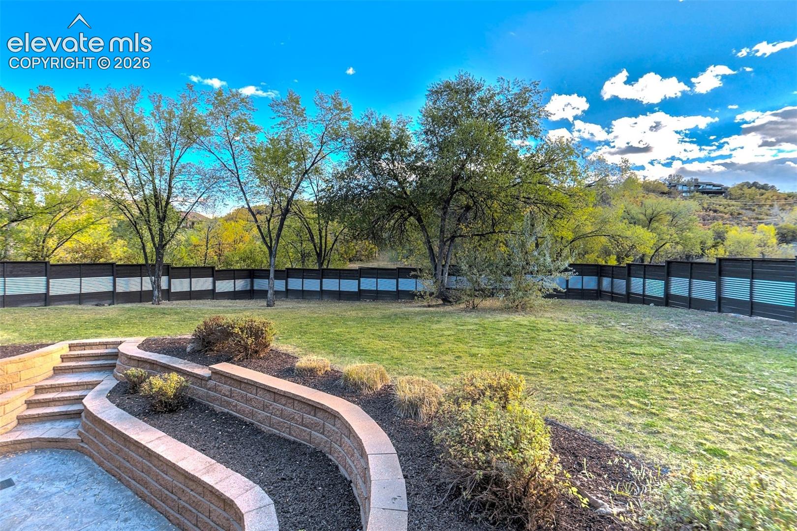 Backyard featuring a tiered stone retaining wall, paved patio area, manicured lawn, perimeter fencing, and mature trees