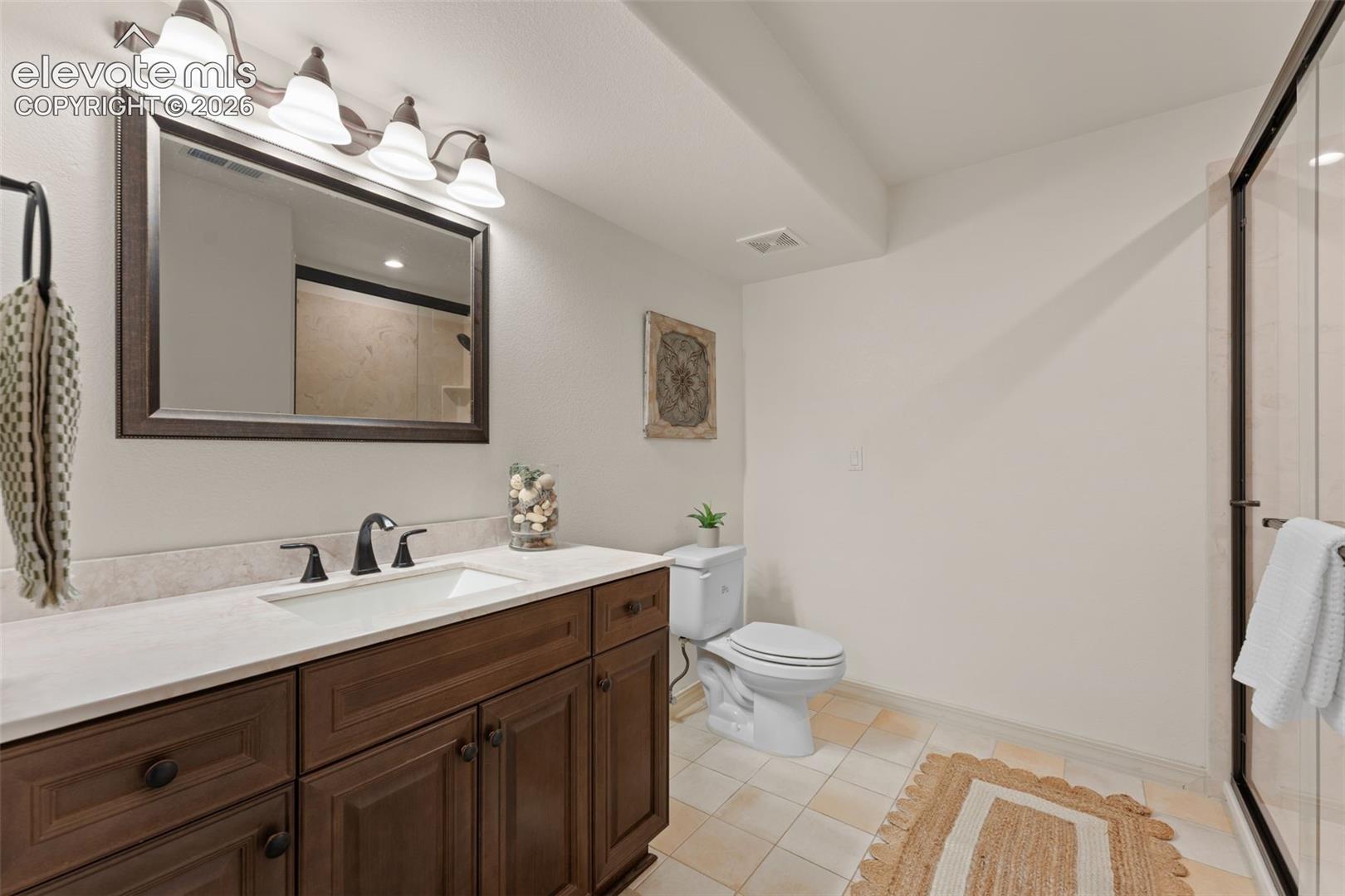 Bathroom featuring a dark wood-finish vanity, integrated countertop with rectangular basin, dark metallic hardware, four-light vanity fixture, and enclosed shower with glass door