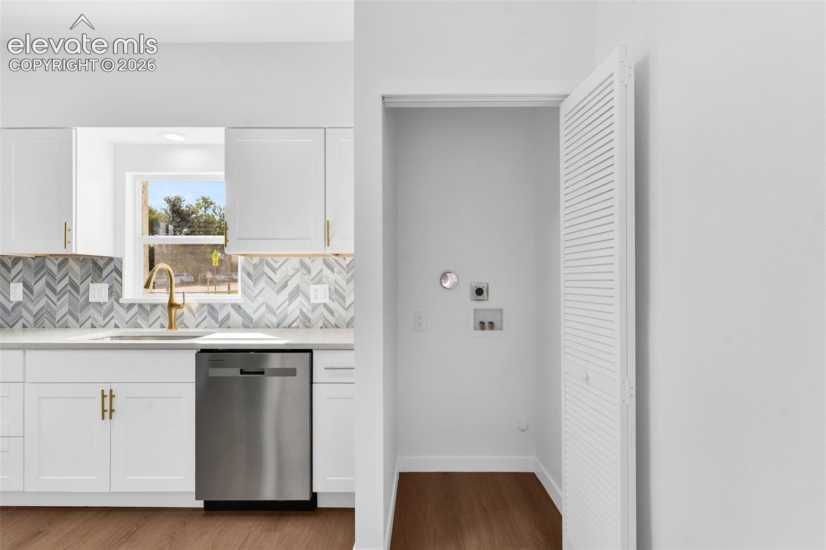 White shaker-style cabinetry with gold-toned hardware, herringbone-patterned tile backsplash, stainless steel dishwasher, brass-finish faucet, and closet with louvered door featuring utility hookups
