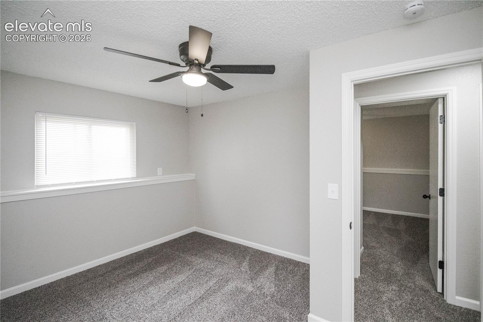 Neutral interior featuring grey carpeting, window with blinds, ceiling fan with integrated lighting, and chair rail molding