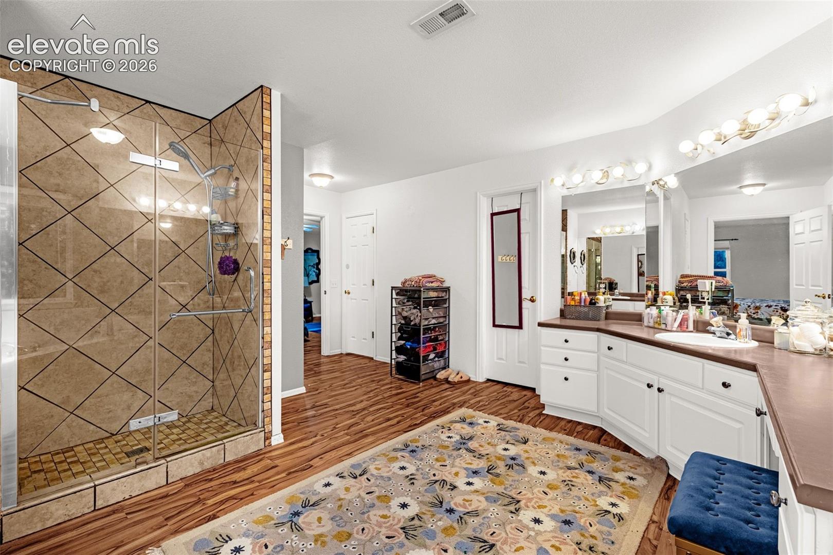 Glass-enclosed shower with diamond-patterned wall tile, wood-finish flooring, white vanity with integrated sink, and large vanity mirror with strip lighting