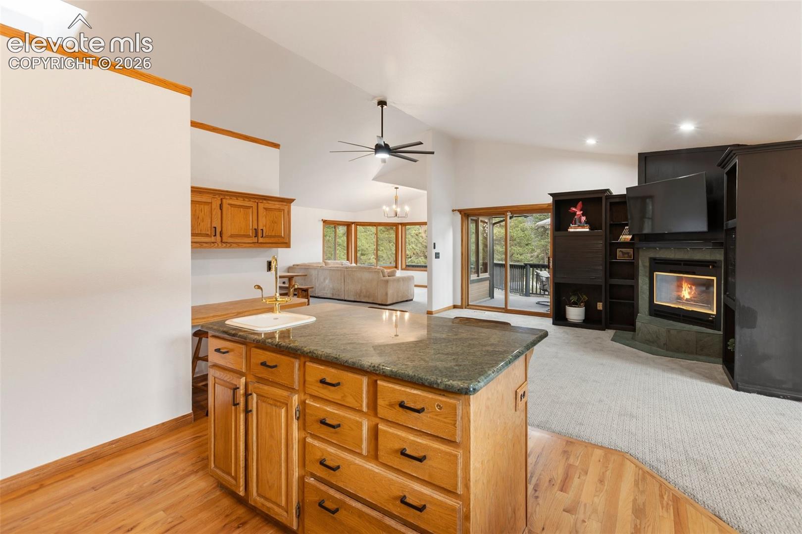 Kitchen island featuring stone countertop, undermount prep sink, and wood cabinetry, vaulted ceiling with integrated lighting, and fireplace with dark-toned built-in shelving