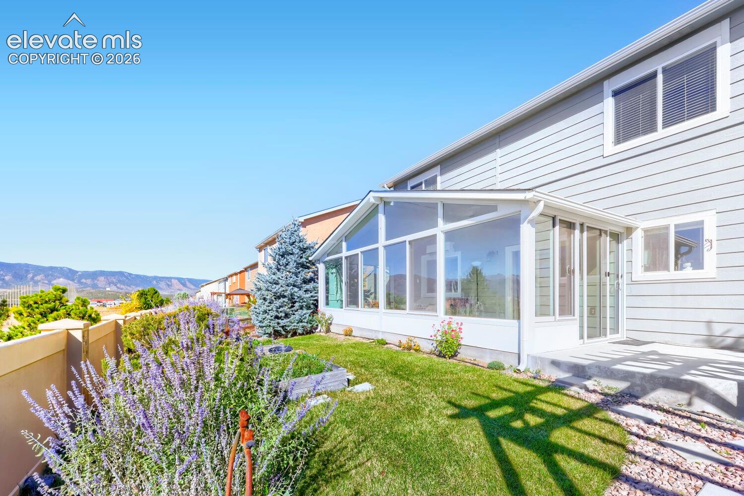 Exterior featuring a glass-enclosed sunroom, horizontal lap siding, a manicured lawn, perennial landscaping, and mountainous scenery