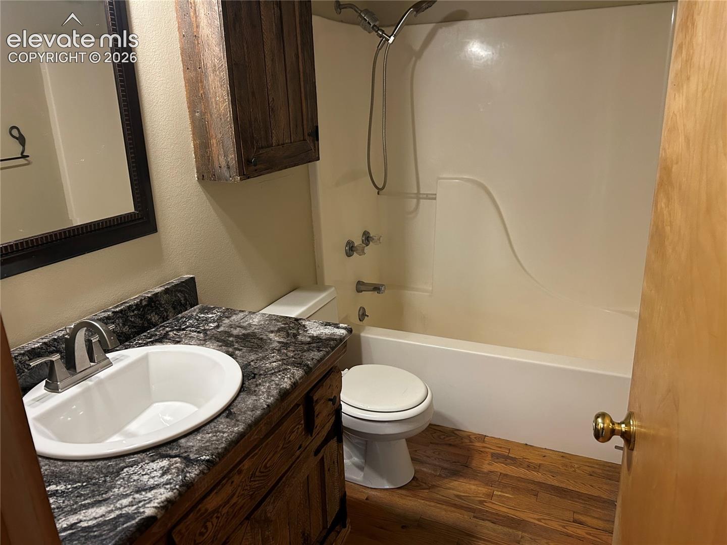 Bathroom featuring wood-finish flooring, a vanity with a dark patterned countertop, an integrated sink, a wall-mounted wooden cabinet, and a tub-shower combination with a handheld fixture