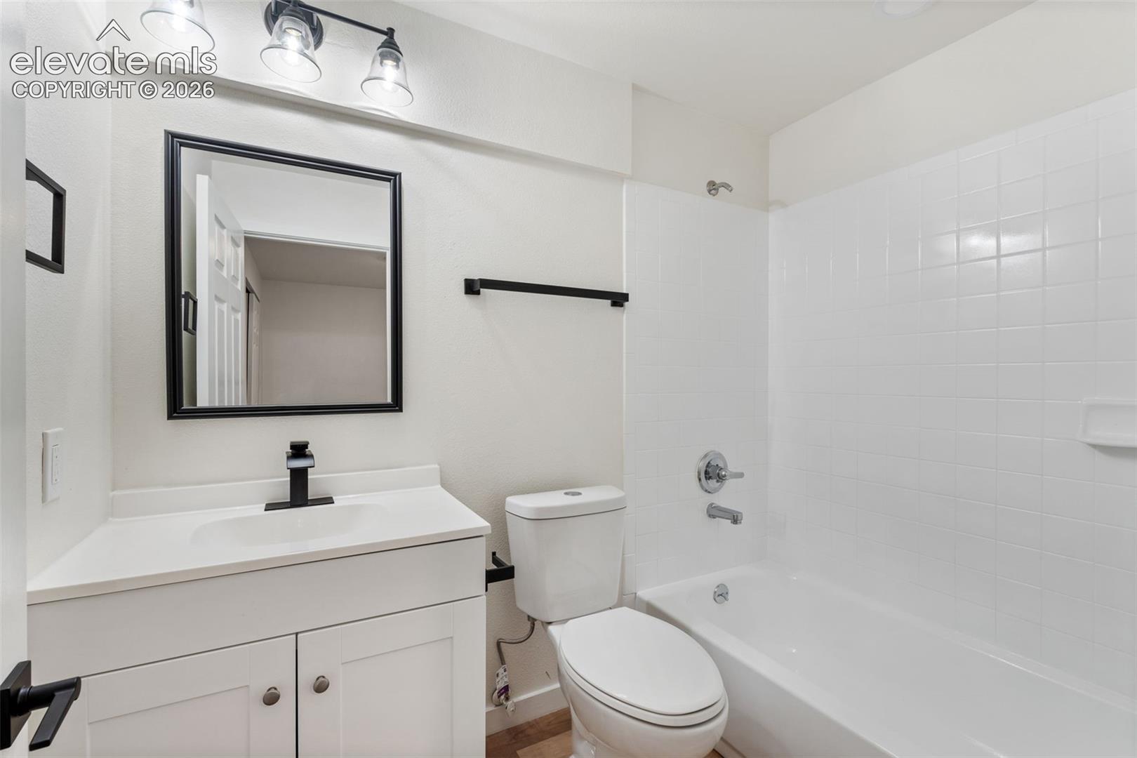 Bathroom vanity featuring a white cabinetry base, integrated countertop, and matte black faucet, complemented by a matching black-framed mirror, three-light fixture, and white tile tub surround