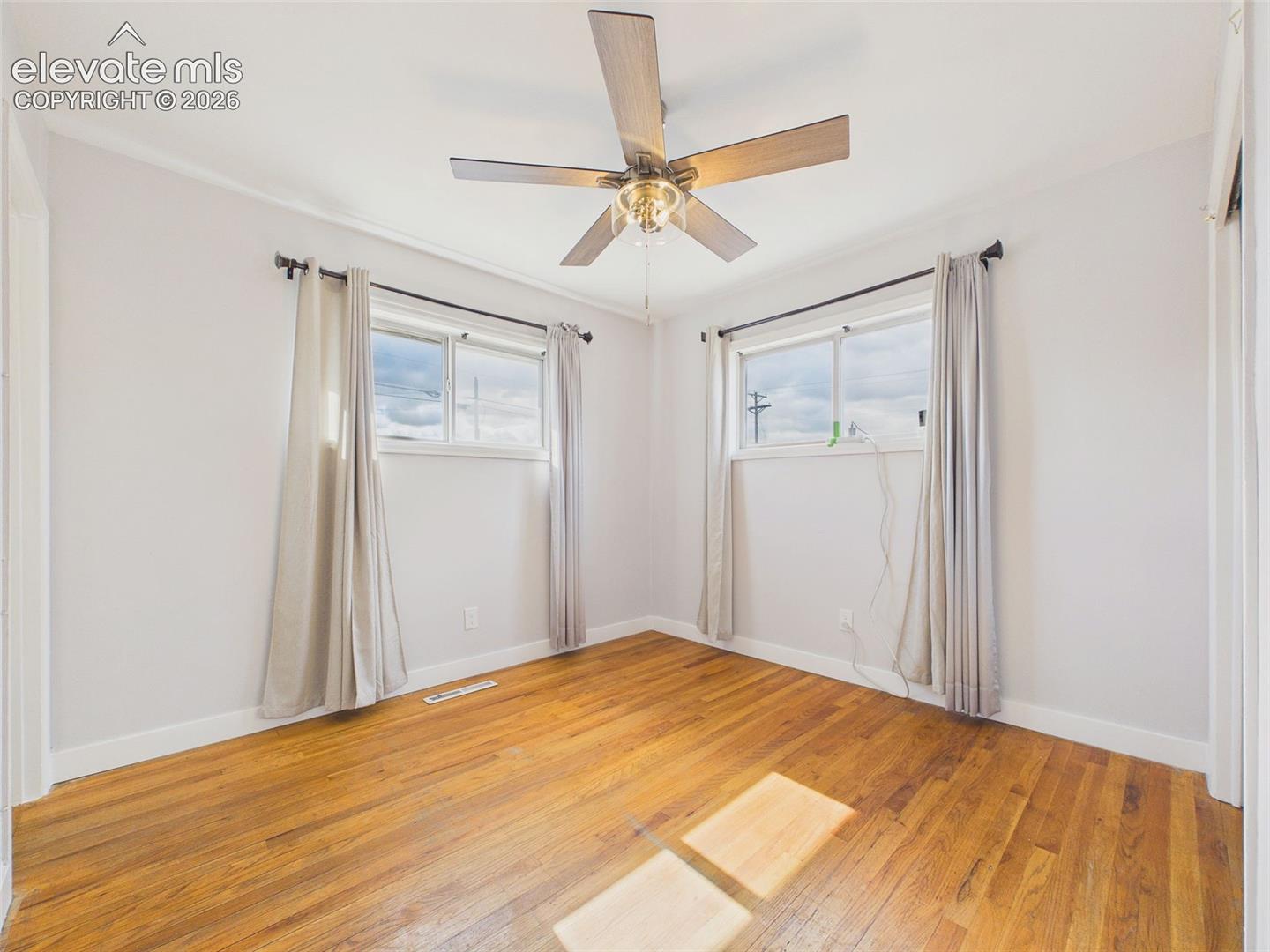 Naturally bright bedroom showcasing hardwood floors, neutral wall colors, two windows, and a ceiling fan with integrated lighting. On suite bathroom