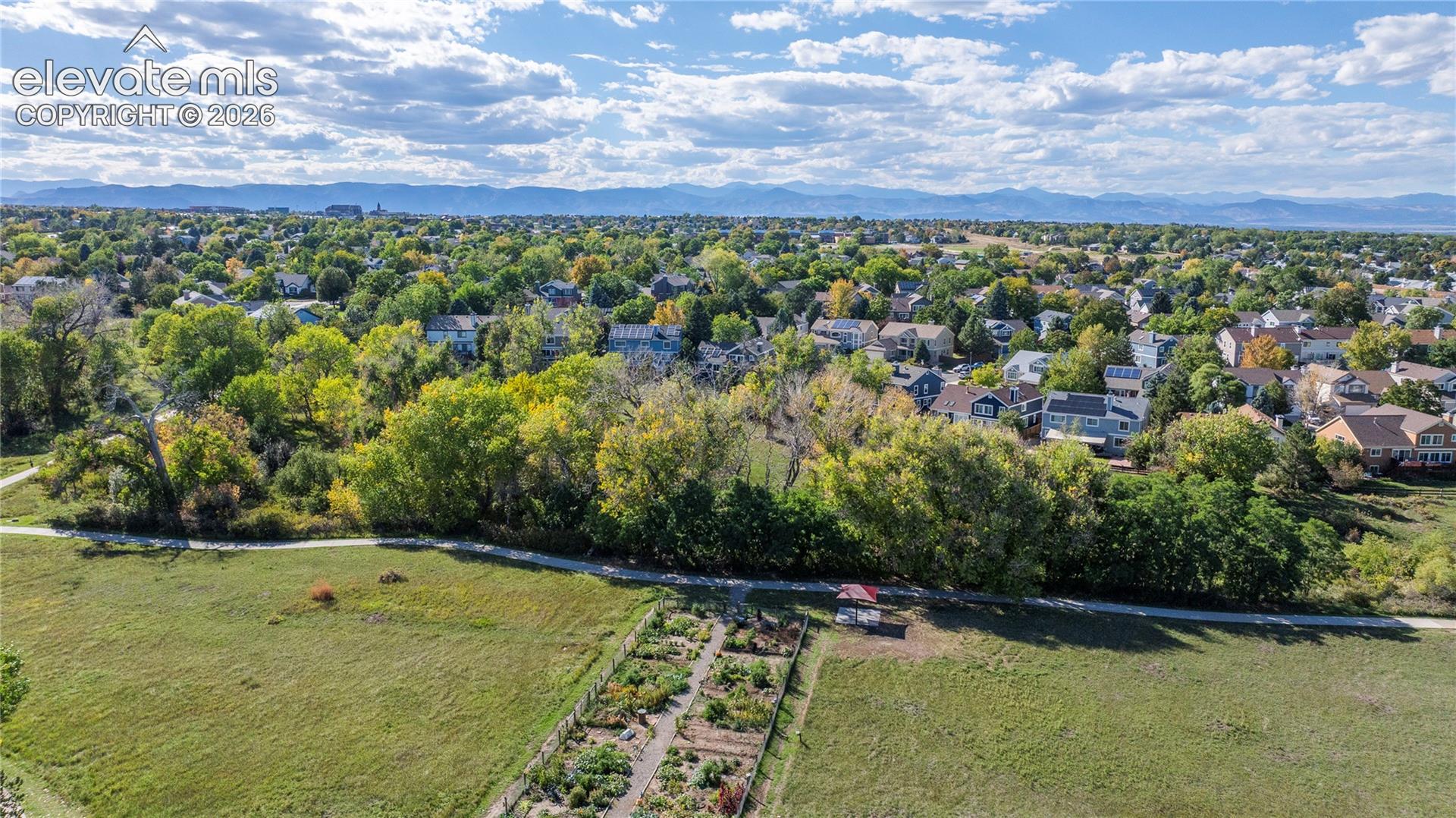 Aerial perspective featuring an expansive open green space, a dedicated garden plot with rows of planting beds, a paved pedestrian trail, mature perimeter trees, and a mountain range backdrop