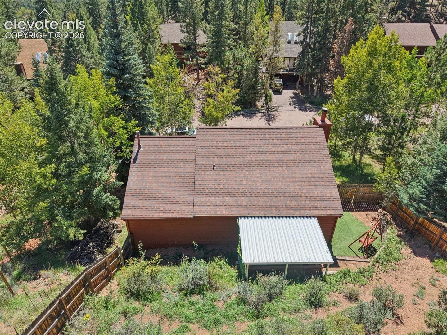 Gable roof with asphalt shingles, red exterior siding, a metal overhang, fenced perimeter, and surrounding mature trees