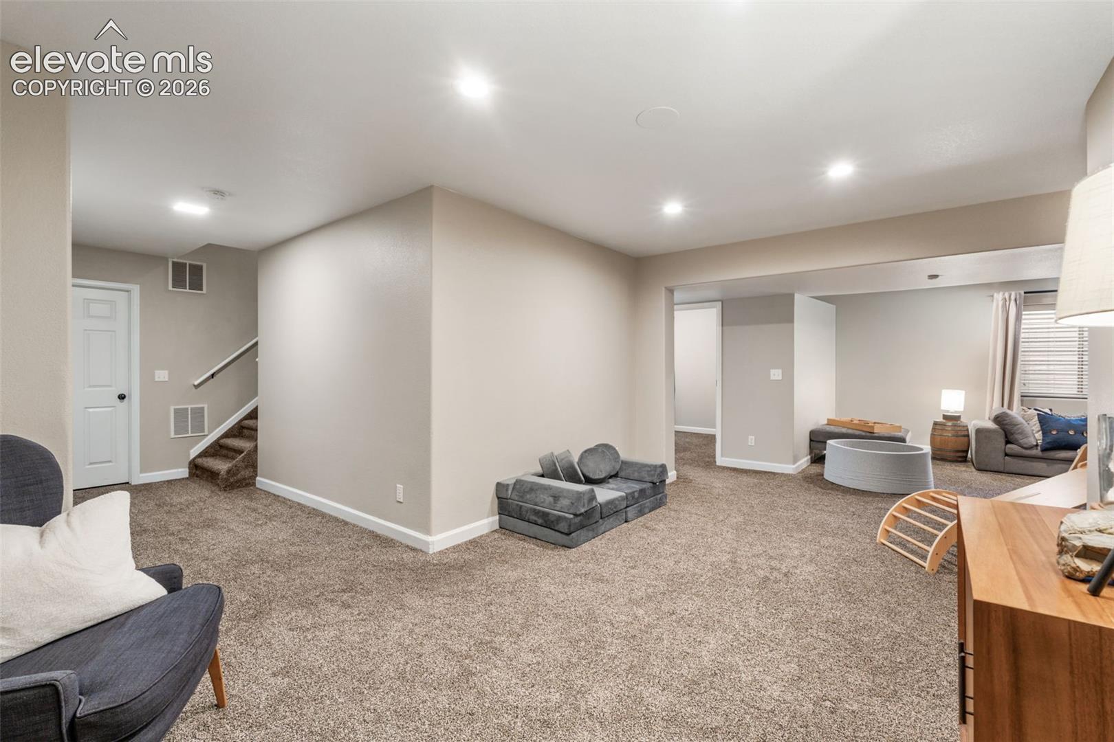 Open-concept interior space featuring neutral wall paint, wall-to-wall carpeting, recessed ceiling lighting, and a staircase with a wooden handrail