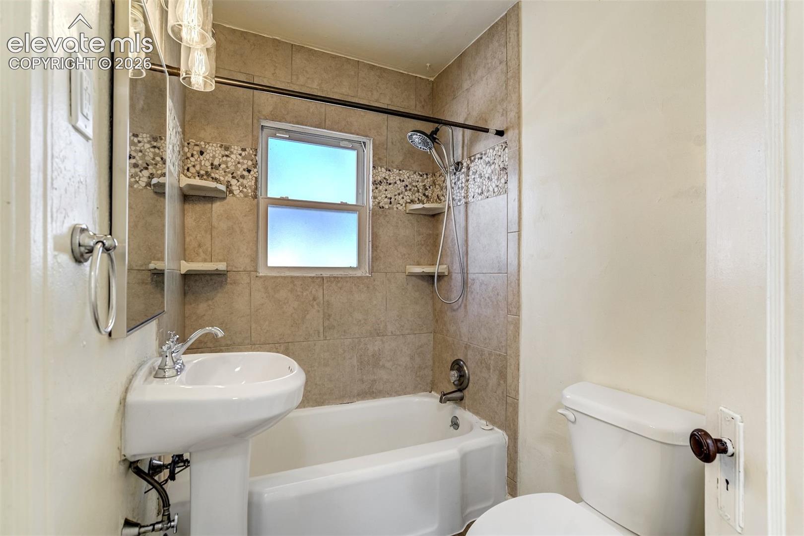 Bathroom featuring a pedestal sink, a bathtub with a tiled surround, integrated pebble-stone decorative inlay, and built-in corner shelving