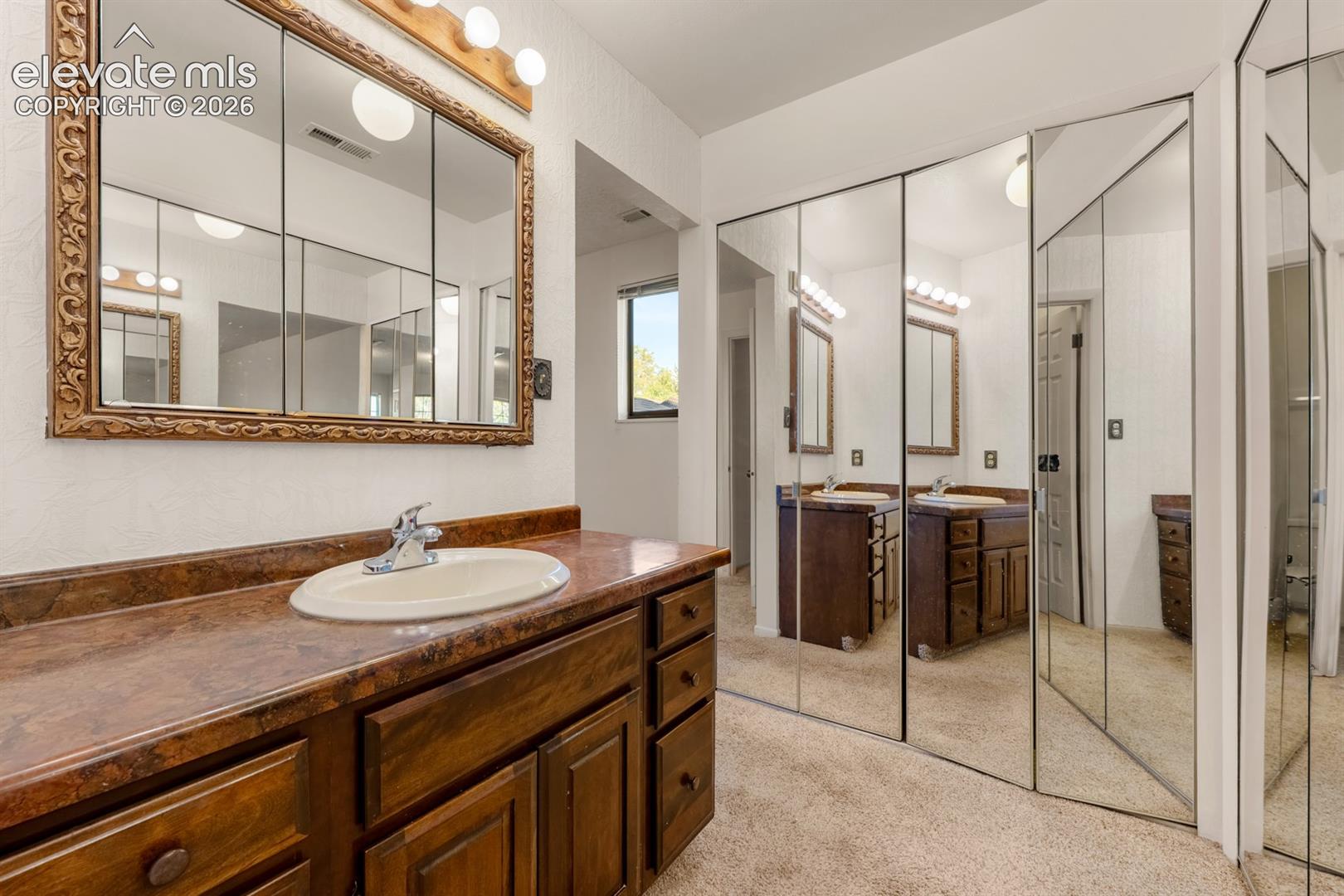 Bathroom vanity with wood-finish cabinetry, solid-surface countertop, integrated oval basin, large framed tri-fold medicine cabinet, and floor-to-ceiling mirrored closet doors