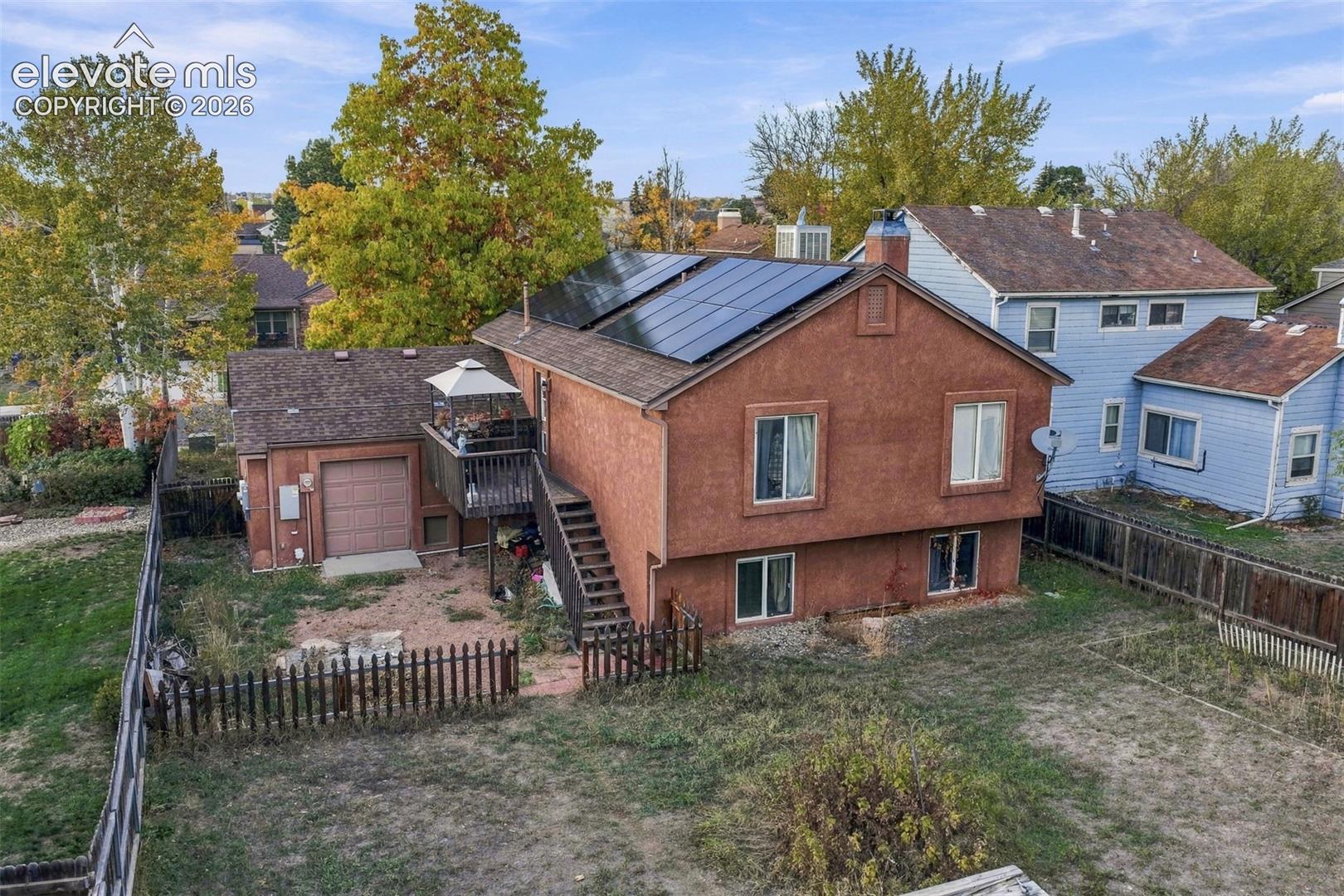 Stucco exterior residence featuring rooftop solar panel arrays, an elevated rear deck with staircase access, and fenced perimeter grounds