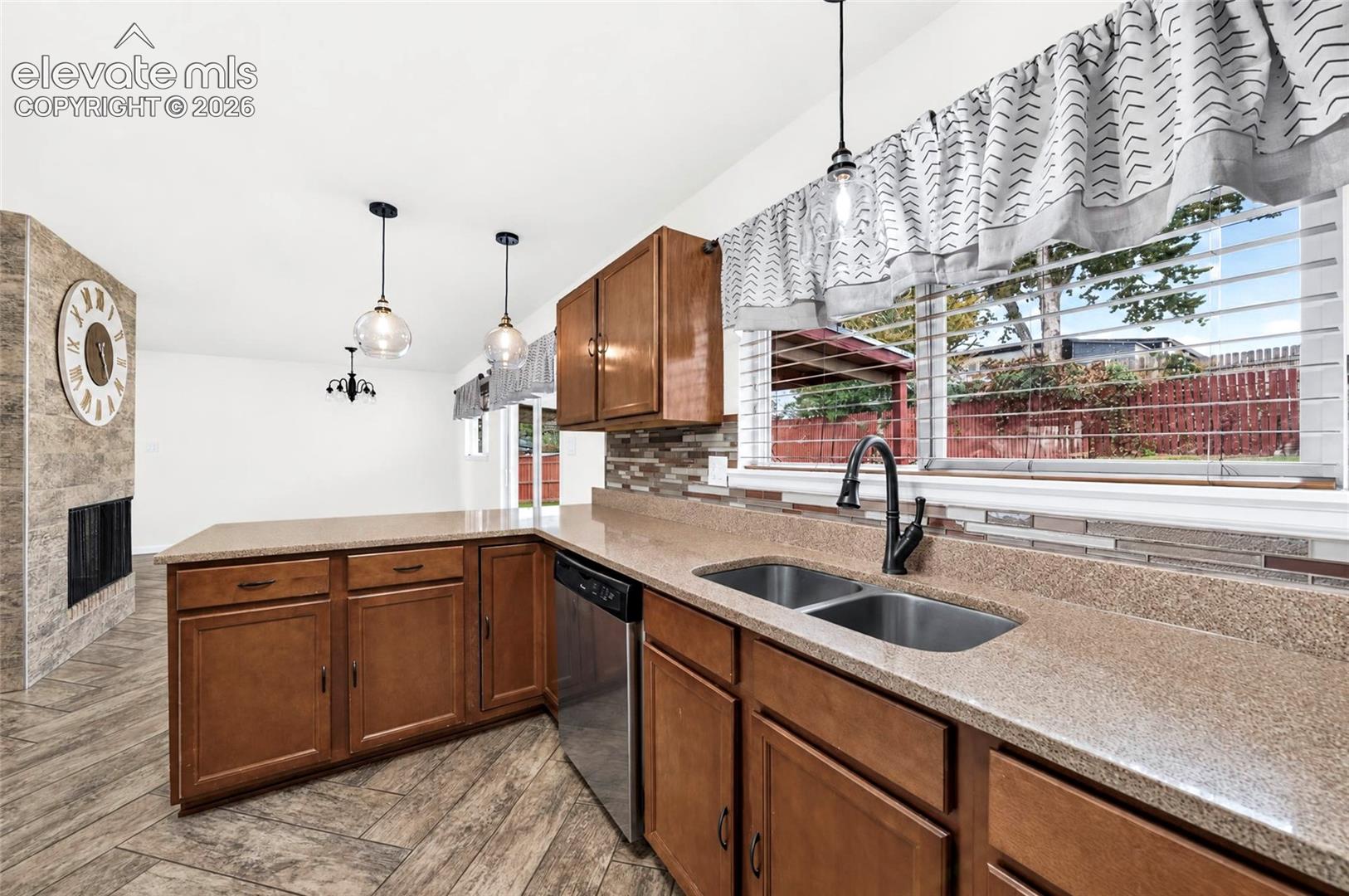 Kitchen featuring wood cabinetry, stone-finish countertops, stainless steel dual-basin sink with matte black faucet, and stone-tiled backsplash, accented by wood-finish flooring and pendant lighting