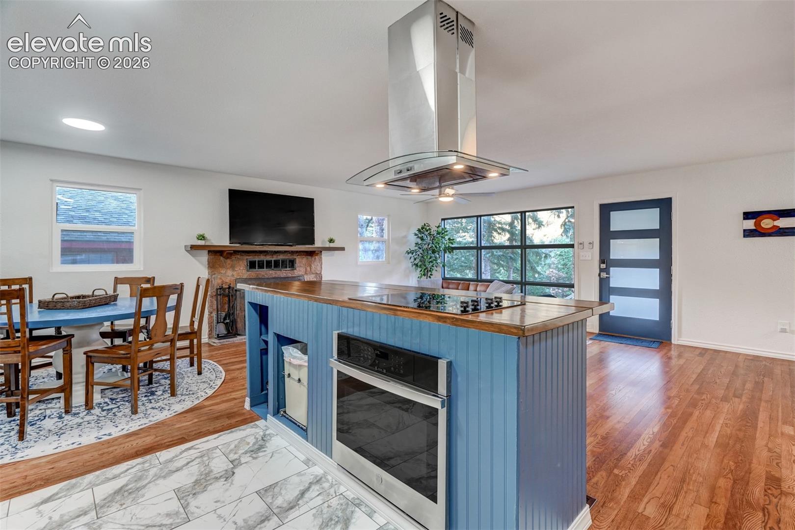 Open-concept living space with hard-surface flooring, a built-in fireplace, modern kitchen island with cooktop and oven, stainless steel range hood, and large multi-pane window